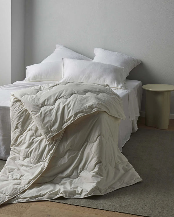 Minimalist bedroom with white pillows and natural linen bedding layered with a quilted wool duvet, soft neutral palette with warm wood flooring and sage accent table