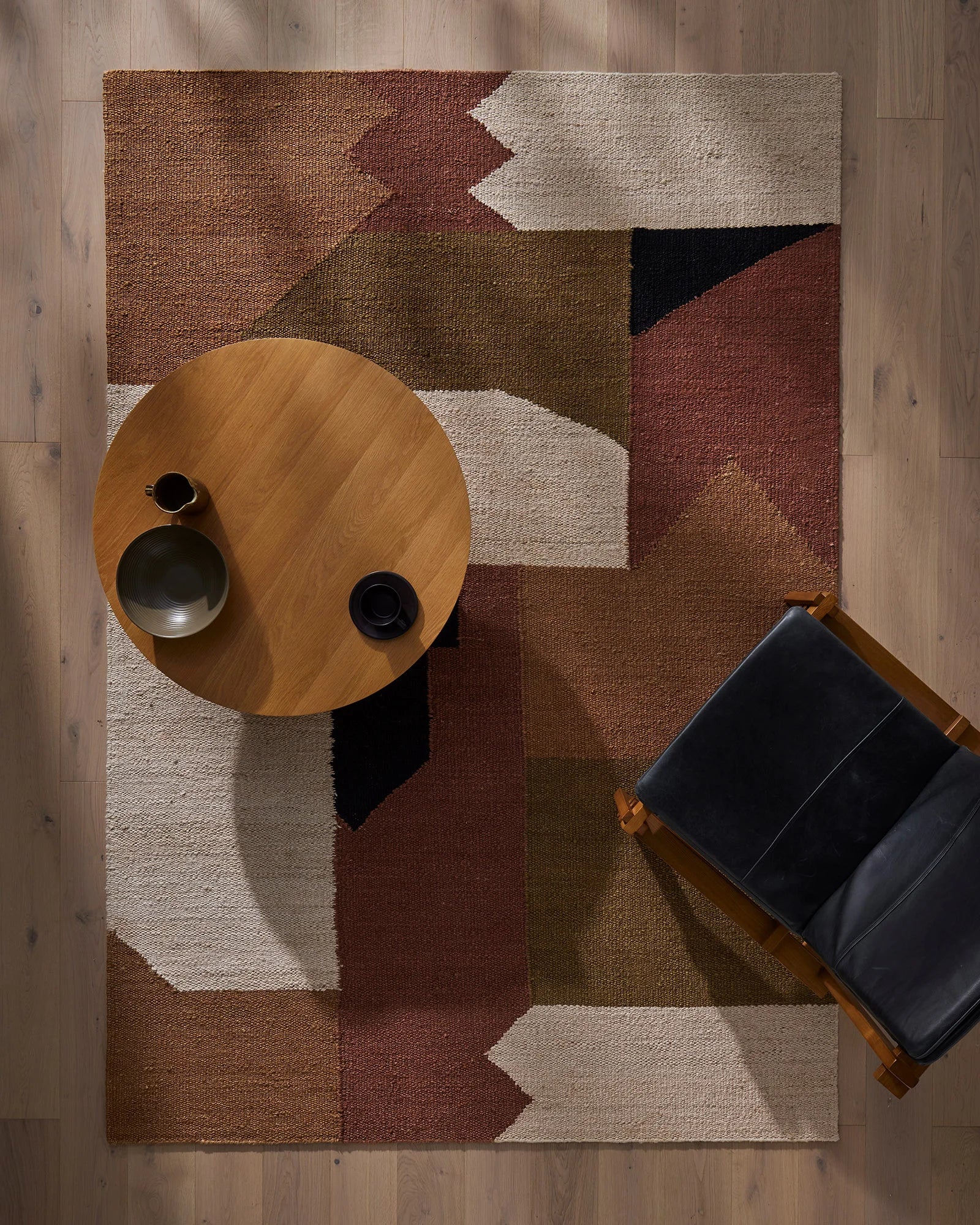 Overhead view of geometric Ruben rug in earth tones—terracotta, olive, cream and charcoal—with timber round table and black leather chair on warm wood flooring