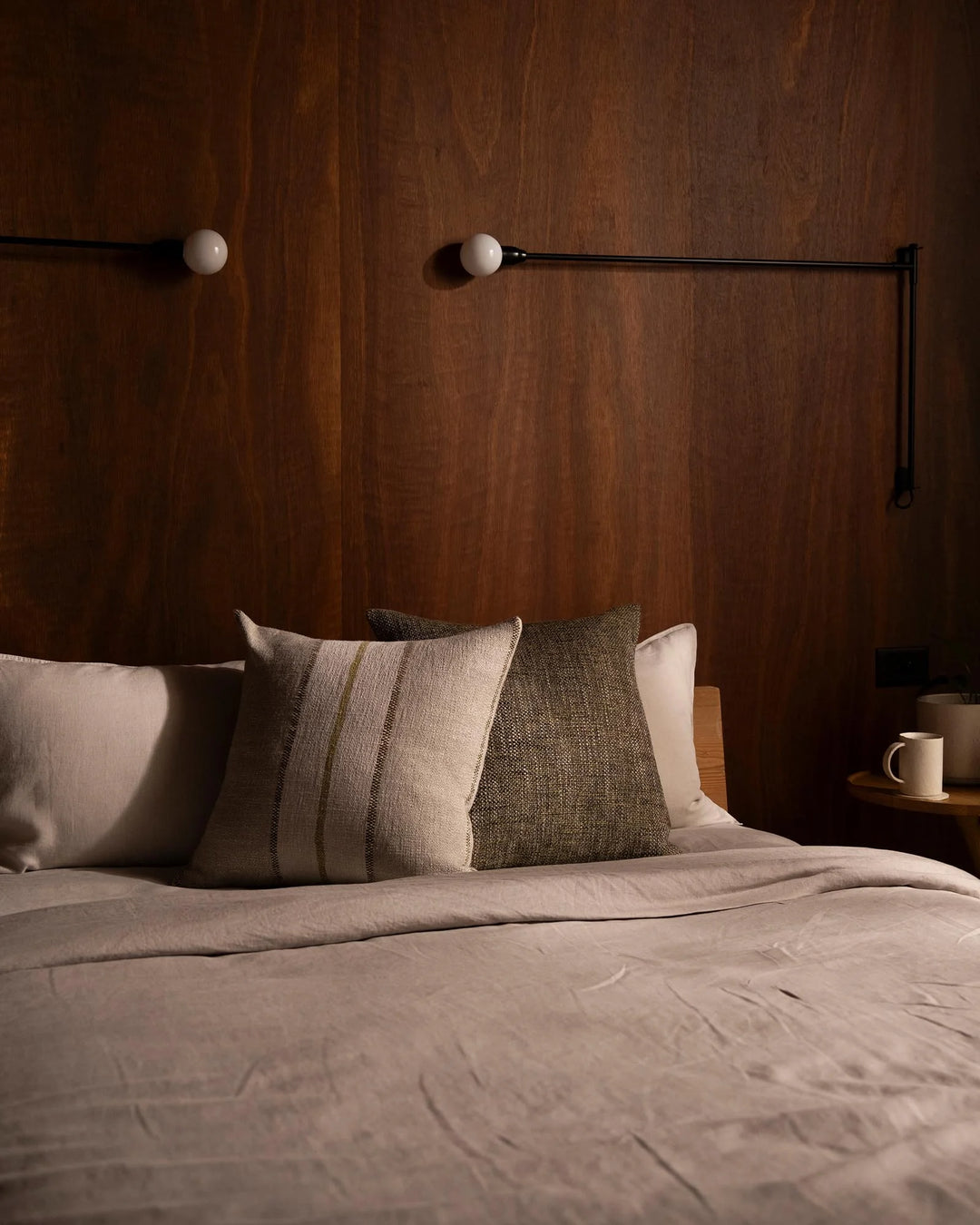 Warm timber bedroom with layered neutral cushions in sage and taupe tones, soft linen bedding, and minimalist black metal wall sconce styling.