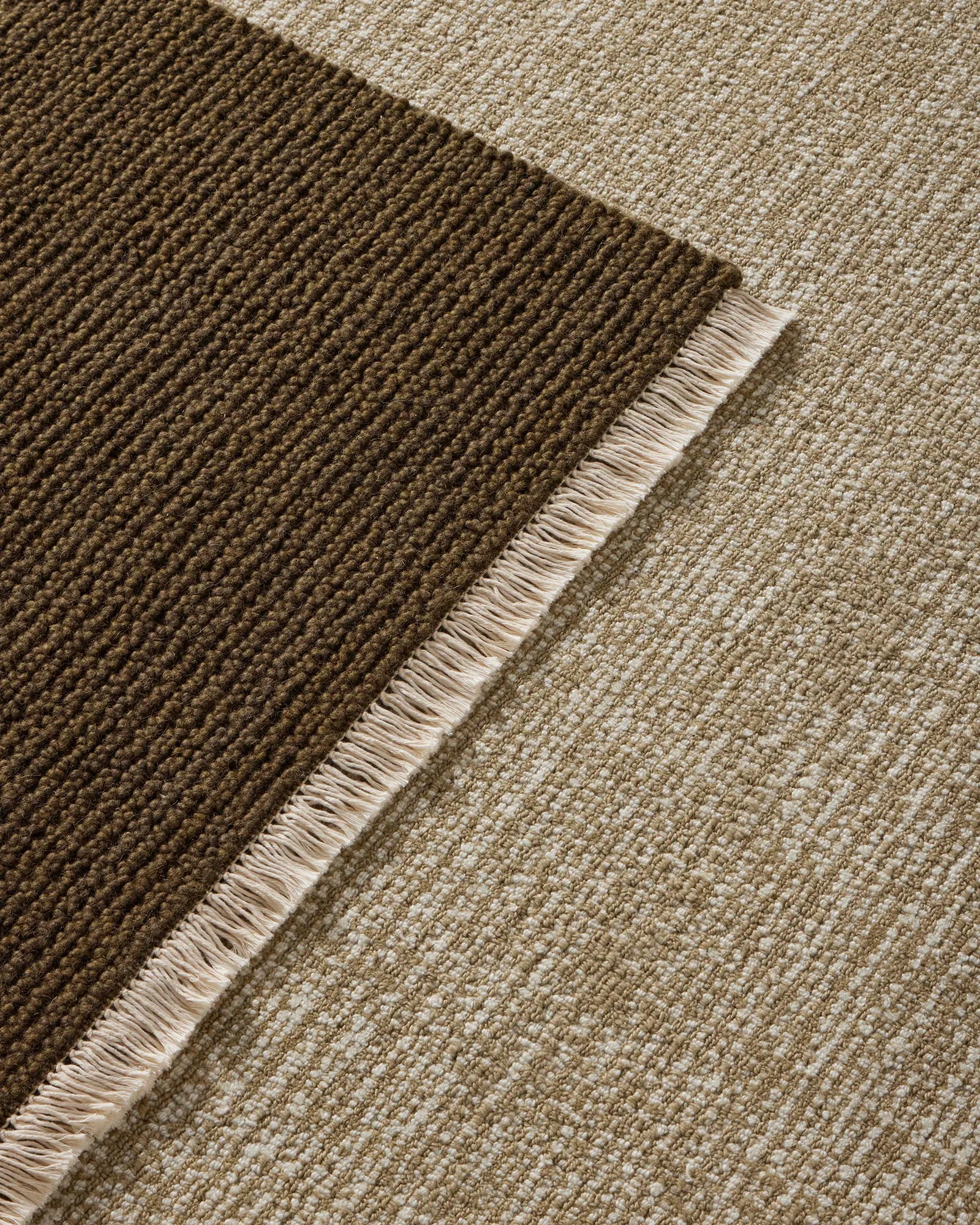 Layered neutral wool rugs in moss and willow tones showcase woven texture and fringed edges, creating warm earthy depth for modern interiors