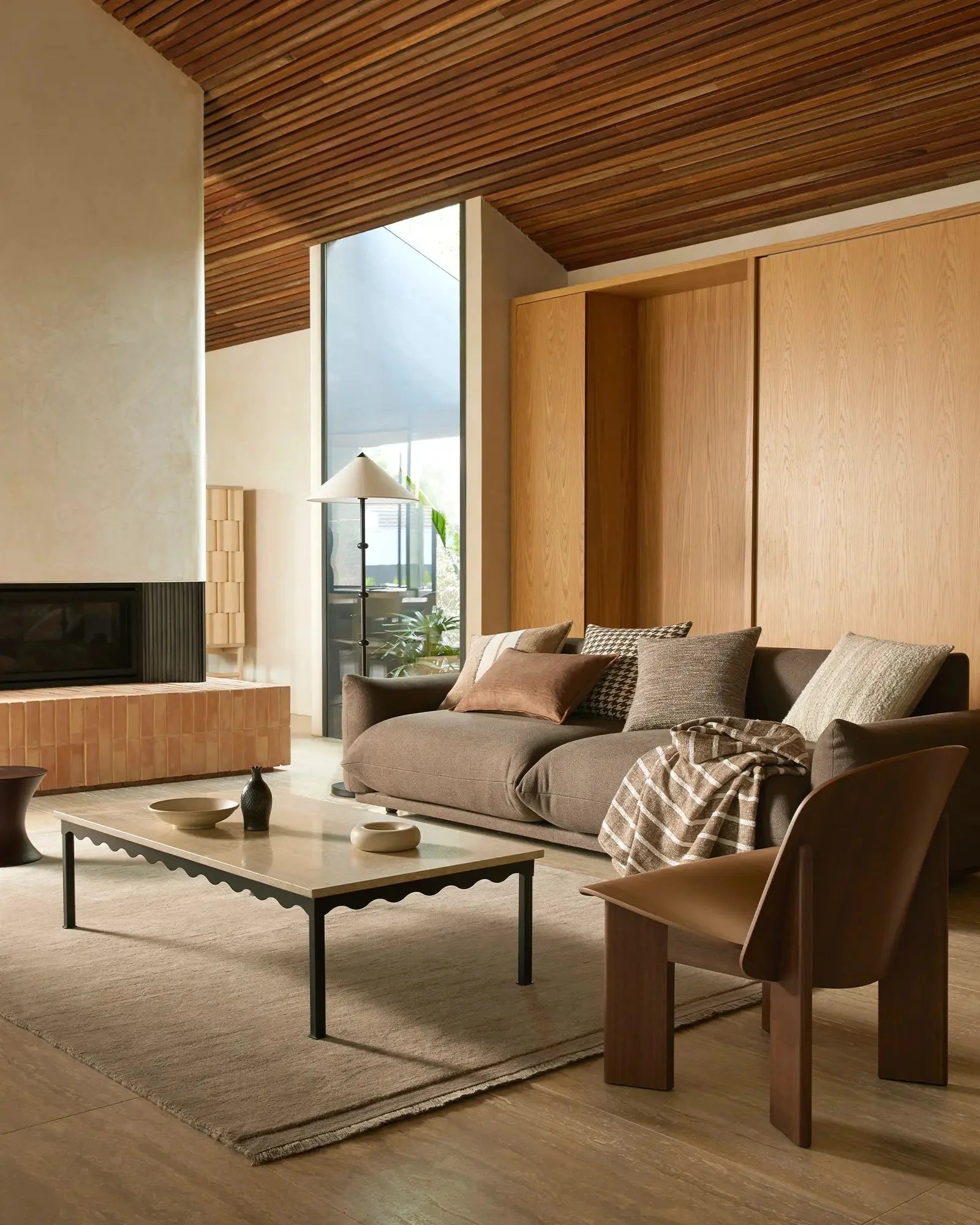 Warm minimalist living room with sloped timber ceiling, terracotta brick fireplace, and neutral earth-toned palette. Piazza throw draped over taupe sofa with Giovanni cushions, wooden furniture, and layered textures in linen, wool, and cotton.