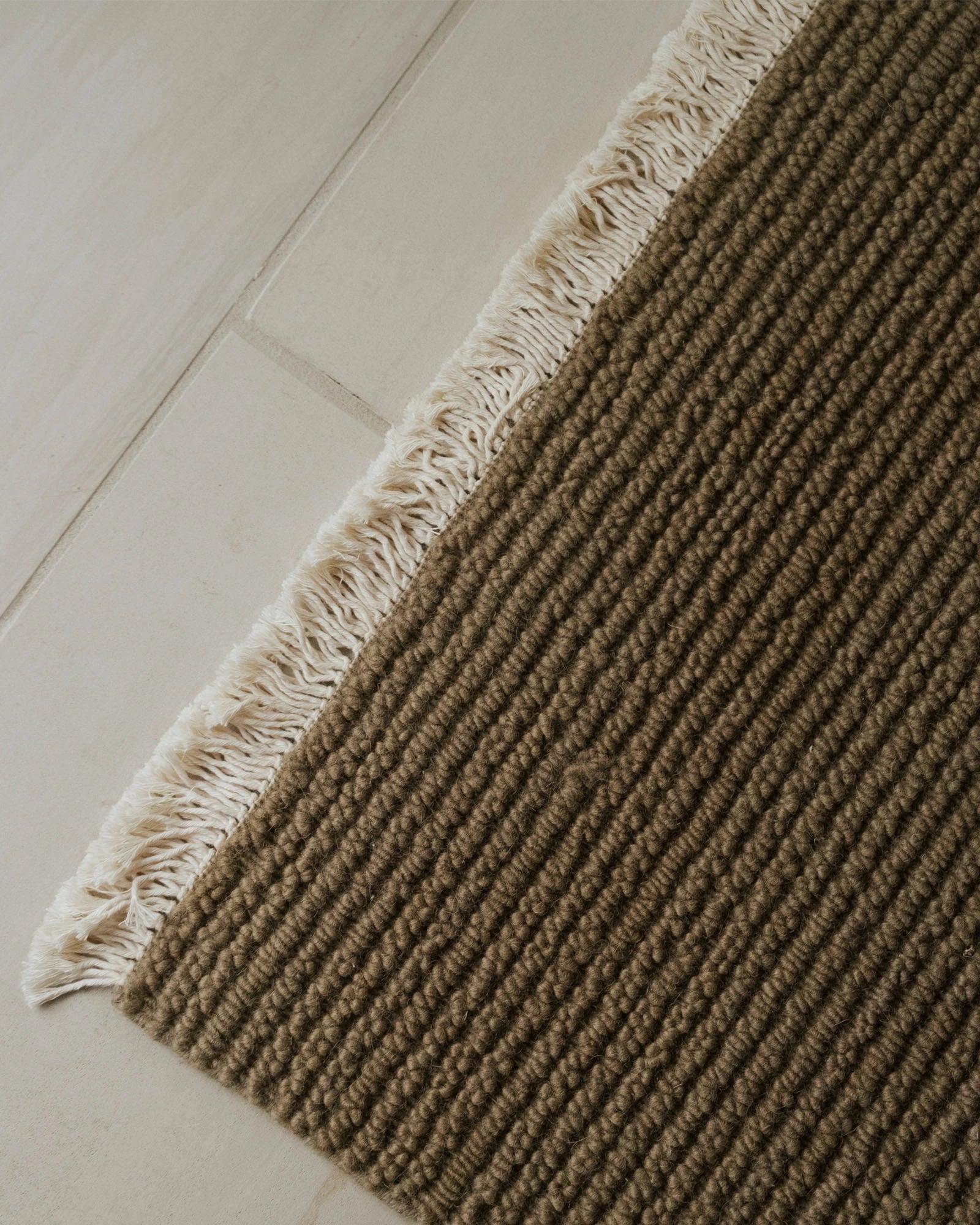 Neutral wool rug with ribbed texture and fringed edge on pale wooden floor, showing earthy taupe and cream tones in minimalist Scandinavian styling
