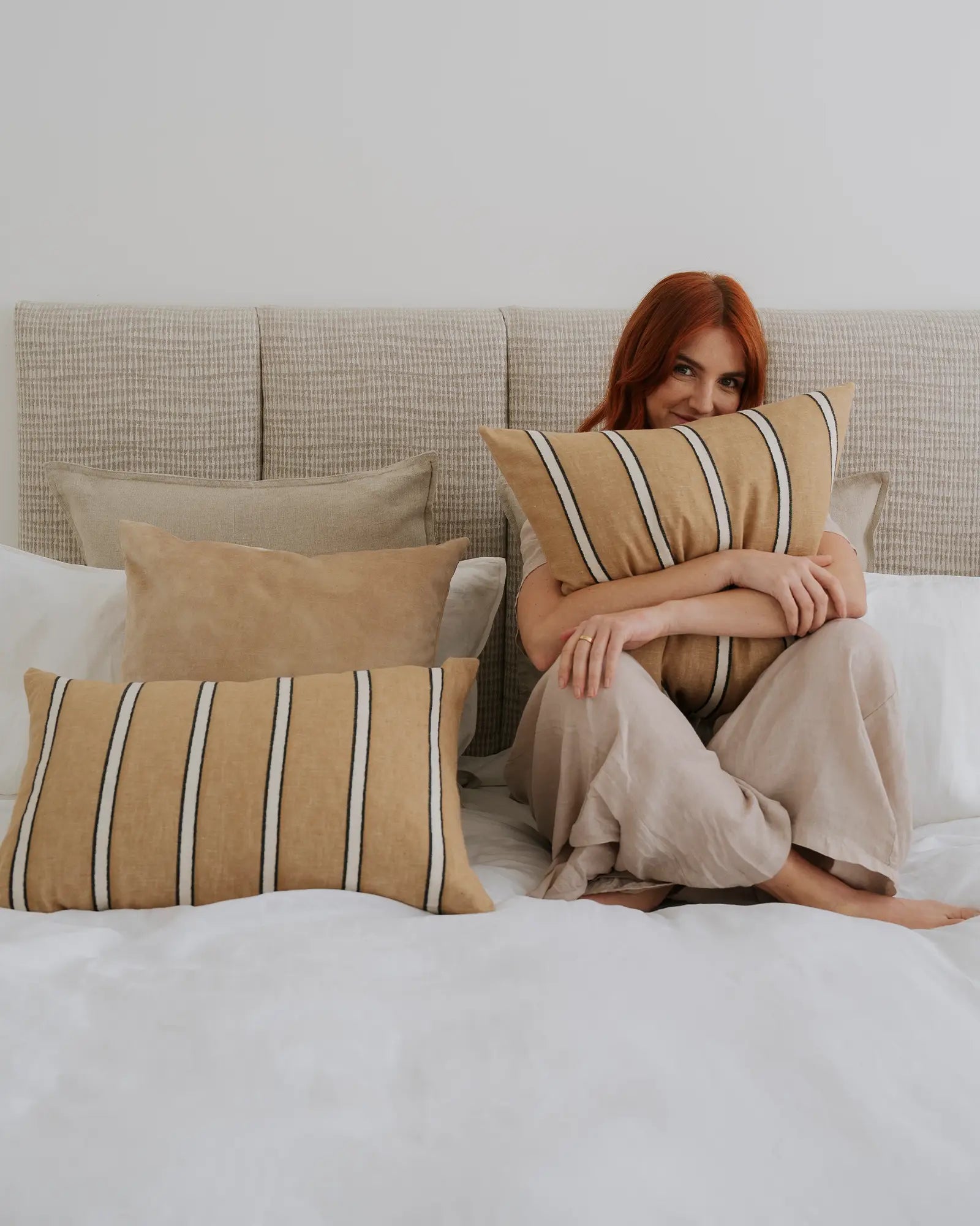 Neutral bedroom styled with warm ochre and charcoal striped cushions layered on natural linen sofa, taupe bedding and white sheets creating a calm, minimalist aesthetic