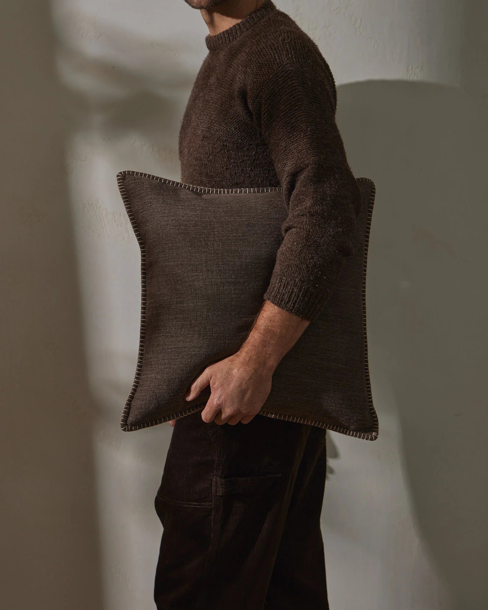 Man in charcoal knit holding a mocha linen cushion with whipstitched edges against cream wall, earthy minimalist styling