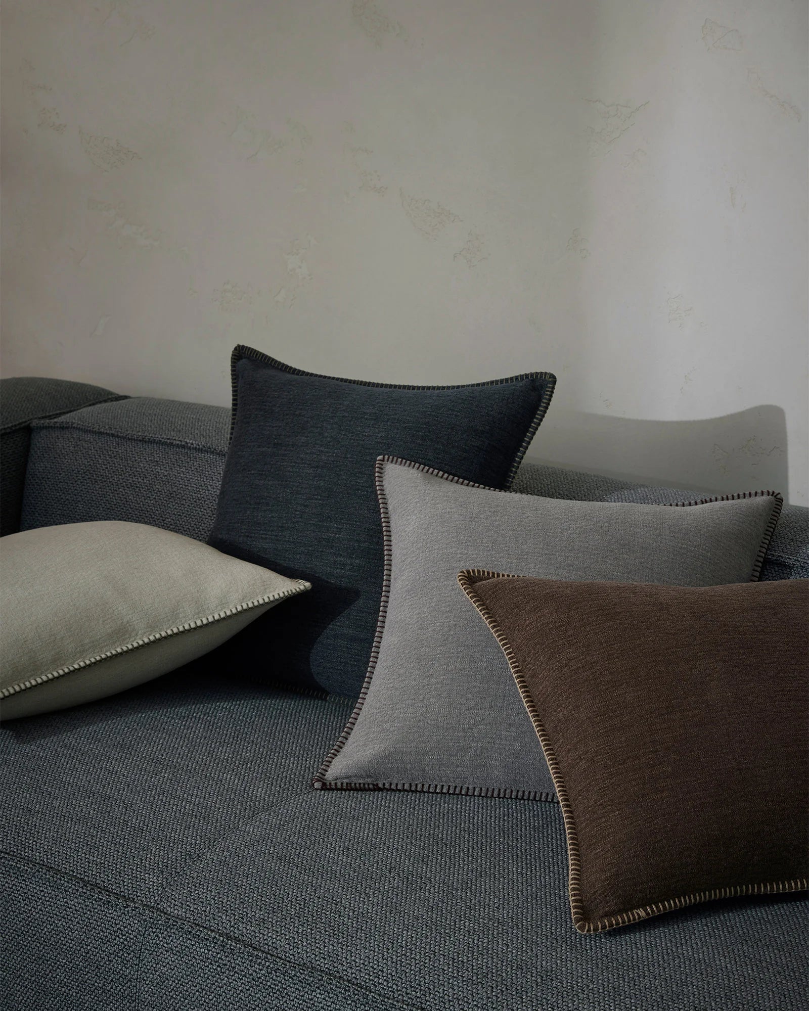 Neutral bedroom styling with Alba cushions layered on a textured linen bed in charcoal, stone and chocolate tones, featuring contrast blanket stitch detailing