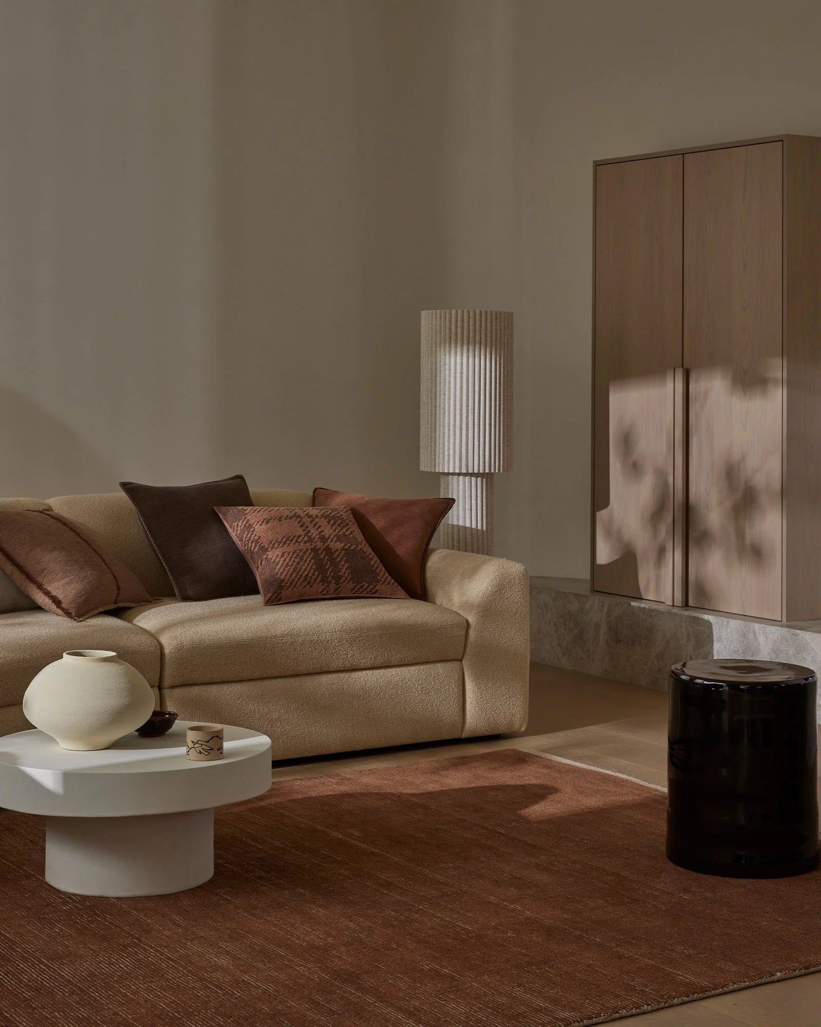 Warm minimalist living room styled with earthy tones, cream linen sofa layered with chocolate and burnt sienna cushions, Remo Amber wool rug, and sculptural ceramic accents