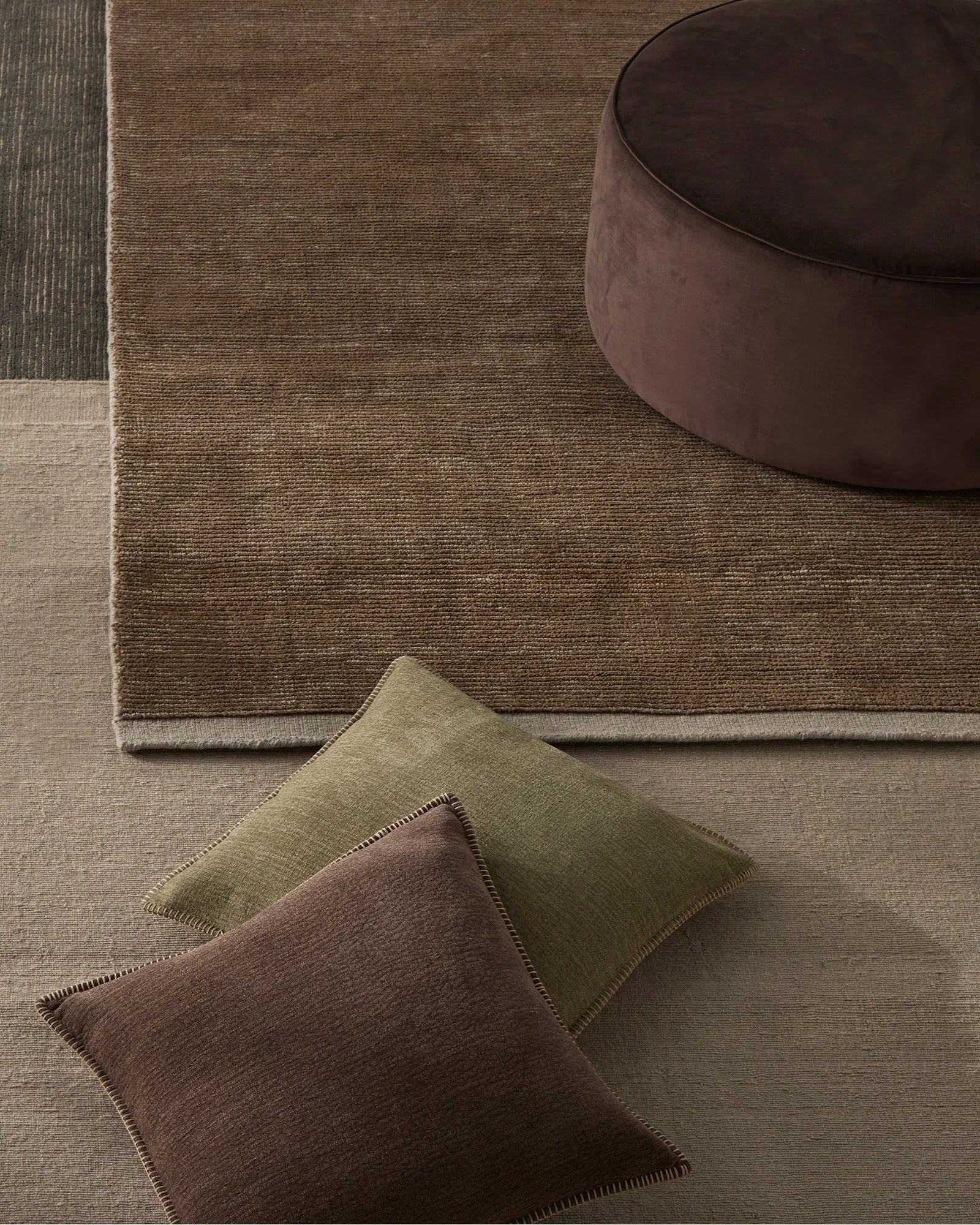 Earthy neutral layered interior with Remo wool rug in warm taupe, Alba cushions in moss and bark tones, and chocolate ottoman on soft linen flooring