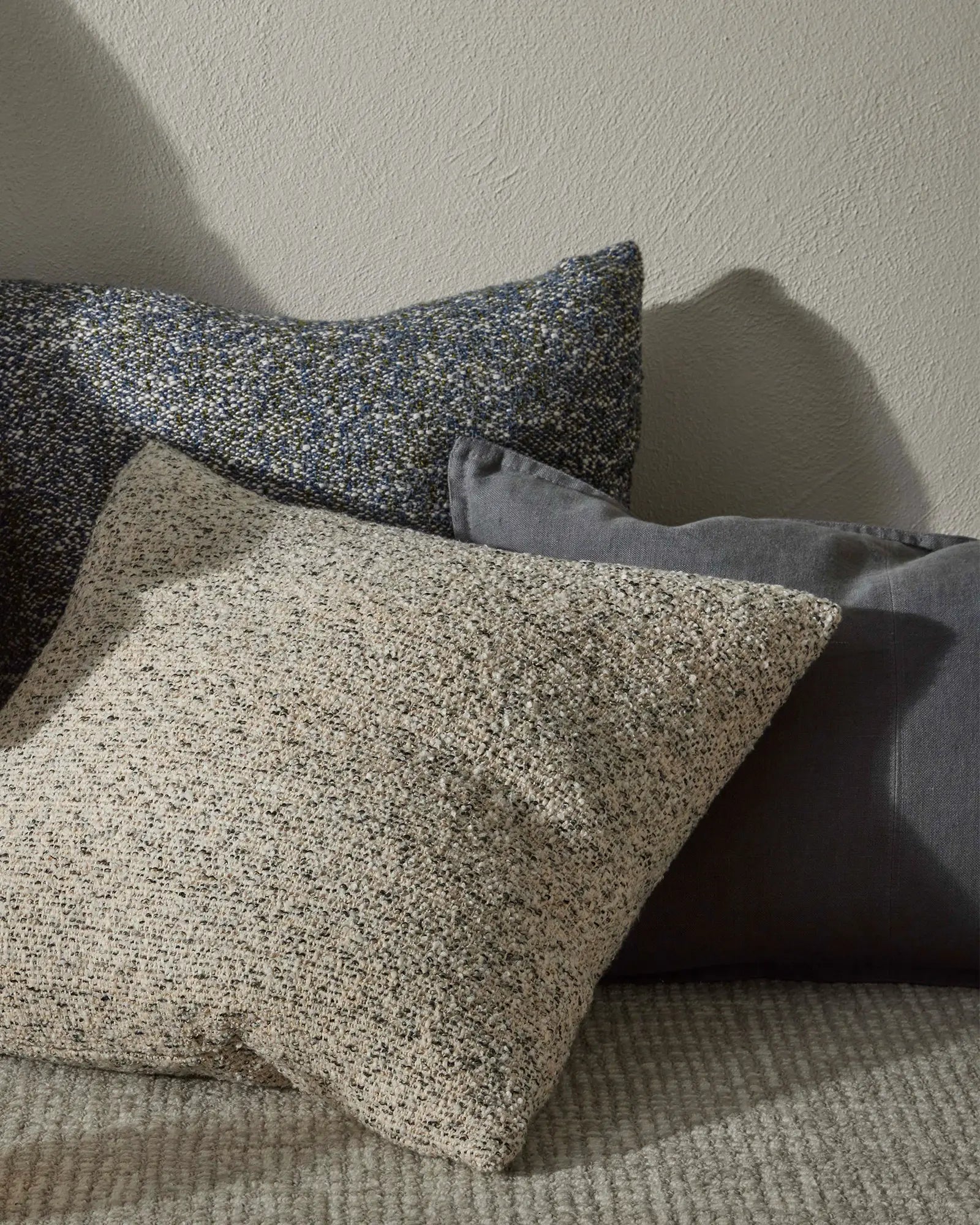 Neutral textured cushions layered on soft furnishings, featuring speckled natural and charcoal tones against pale walls. Minimalist styling with tactile woven fabrics in earthy, monochromatic palette.