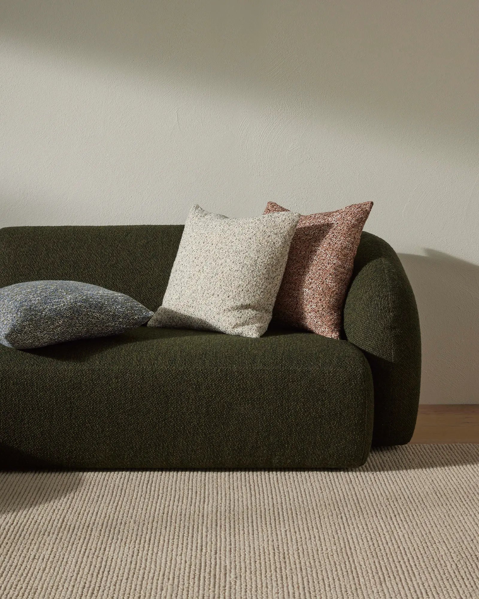 Earthy minimalist living room with sage green curved sofa, cream and terracotta textured cushions, neutral linen rug, and soft natural light creating warm neutral palette aesthetic