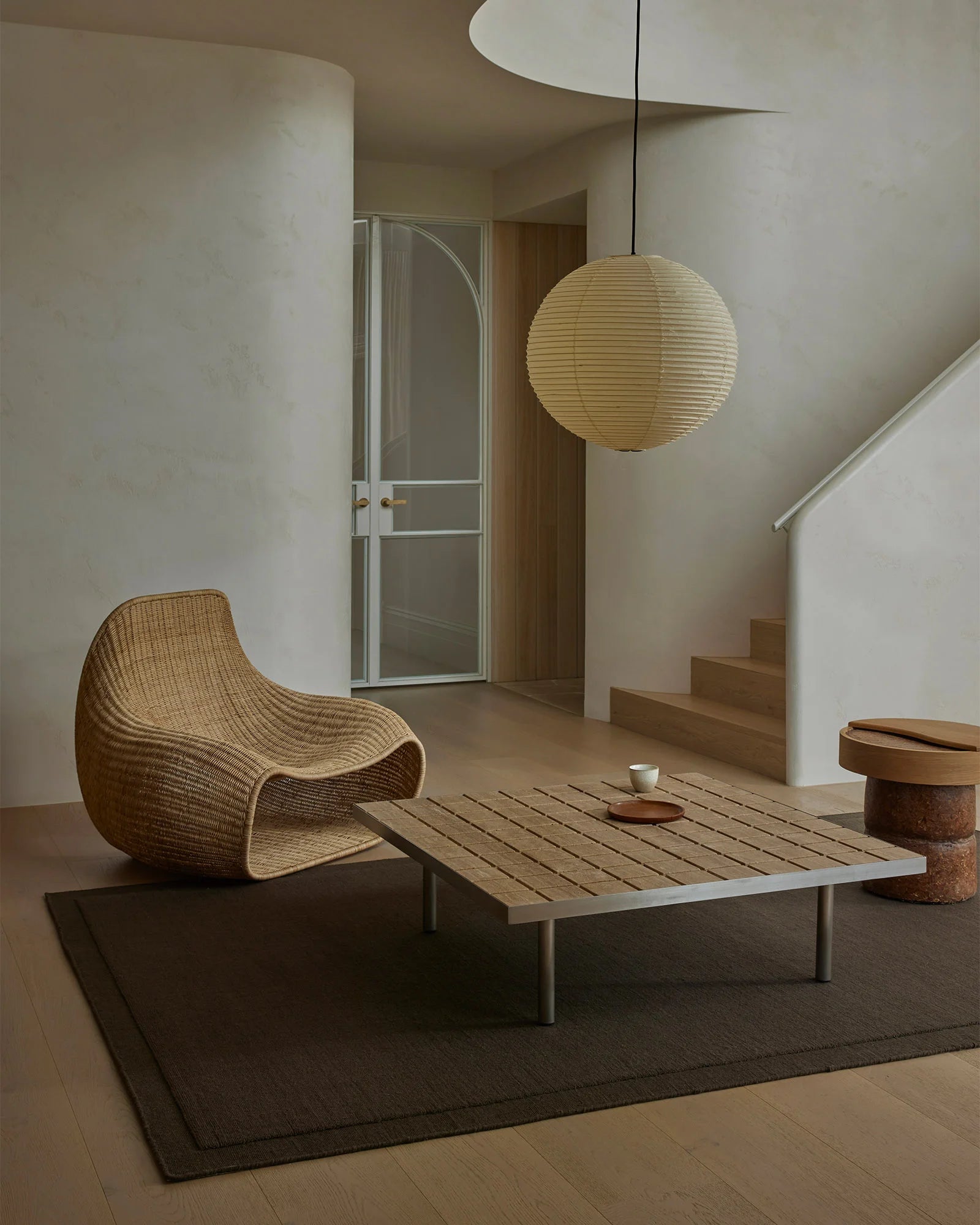 Minimalist living space with natural wood furnishings, woven rattan chair, and charcoal wool rug anchoring the seating area beneath pendant lighting