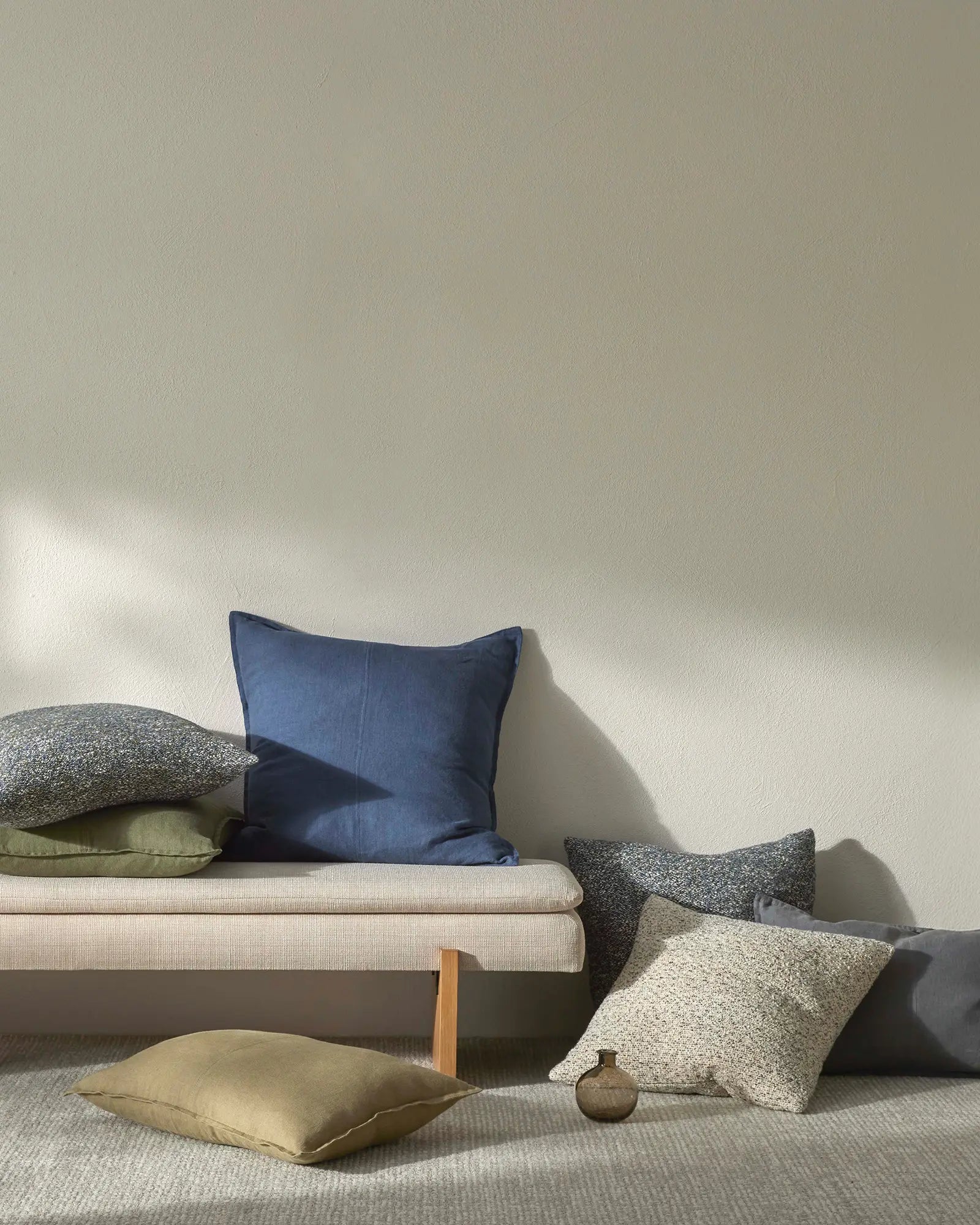 Minimalist living room styled with neutral textiles: cream linen sofa, sage and charcoal linen cushions, ochre silk pillow, and soft grey wool rug against white walls with natural light