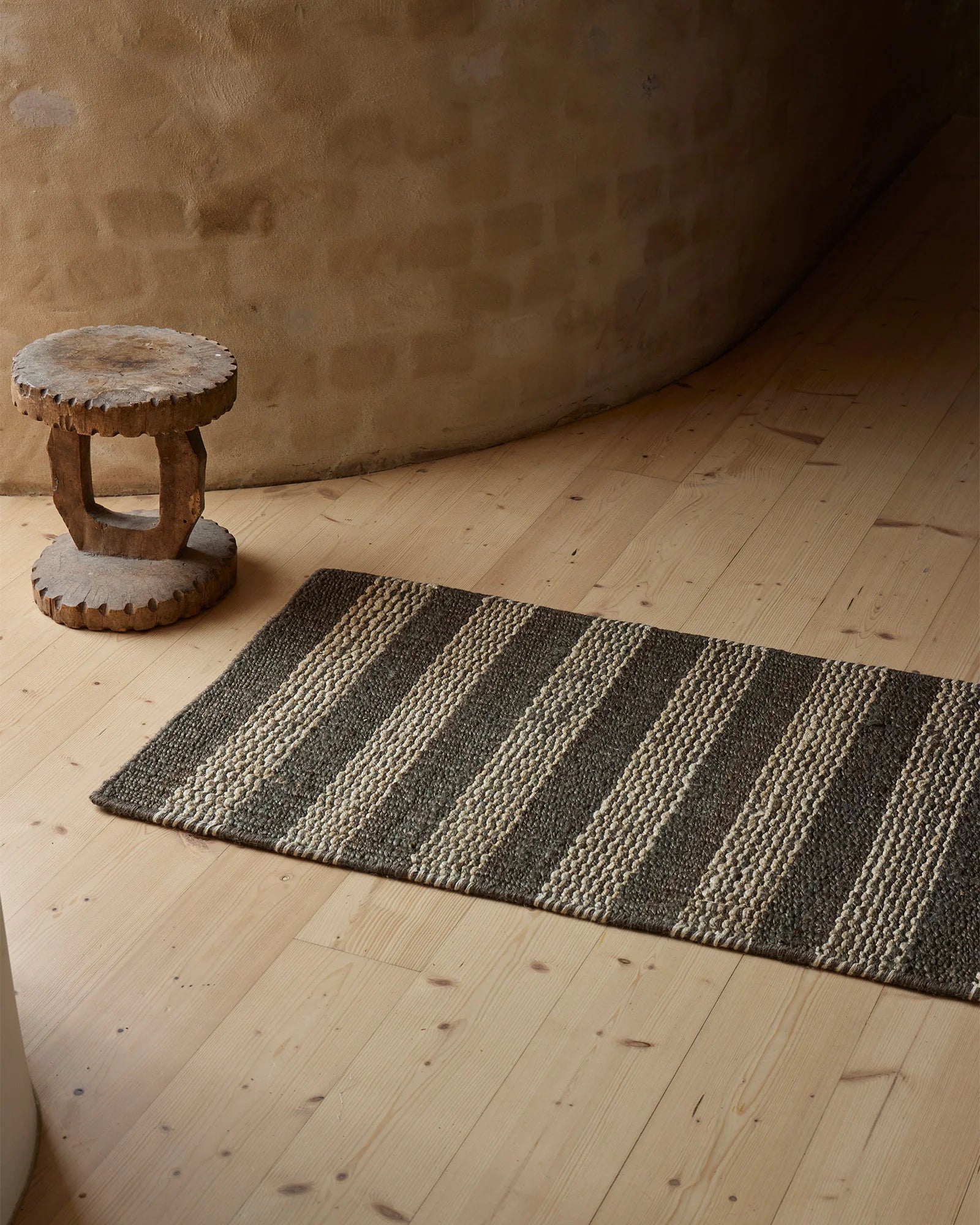 Charcoal and cream striped jute doormat styled on light timber flooring with carved wooden stool and textured plaster wall, showcasing neutral earth-tone interior aesthetic