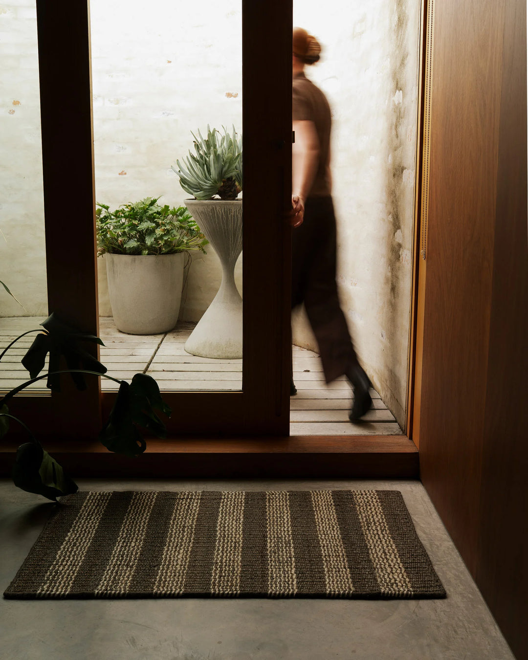 Striped jute door mat in charcoal and cream at entrance with industrial architectural framing, weathered walls, and potted succulents beyond