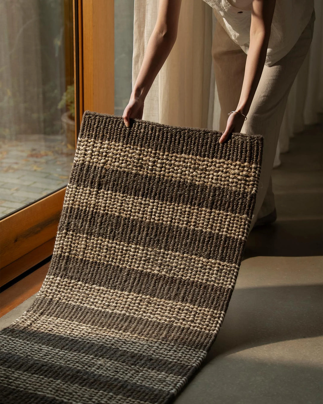 Hands holding charcoal and natural jute door mat in minimalist hallway with warm timber frame and neutral linen curtains, textured weave detail