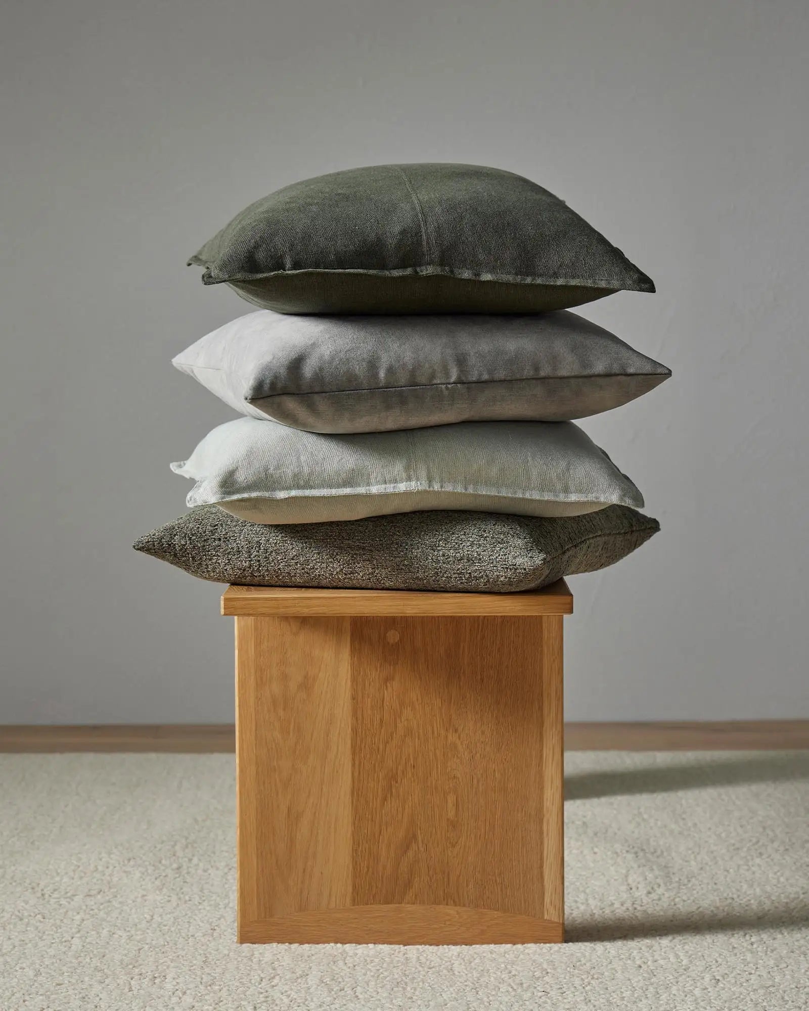 Stacked linen cushions in sage, cream and charcoal tones with textured weaves, displayed on natural timber base against neutral backdrop