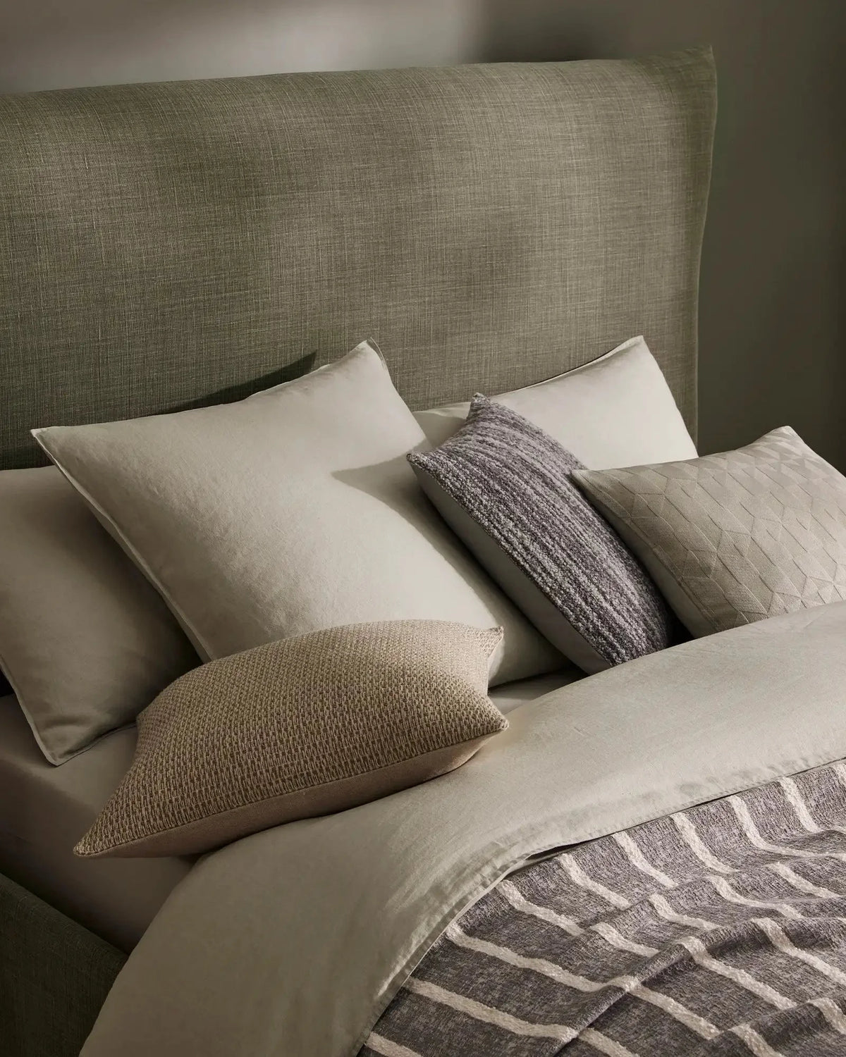 Neutral bedroom scene with layered linen bedding, sage headboard, natural texture cushions in cream and charcoal, and geometric patterned throw in warm grey tones