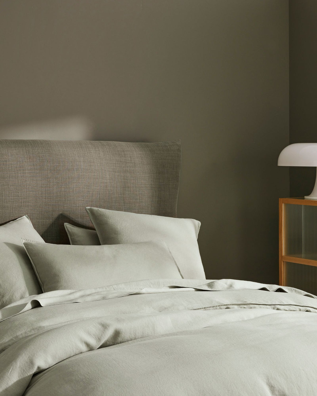 Sage green bedroom with linen Ravello bedding layered in soft naturals, textured upholstered headboard, wooden nightstand and pendant lamp creating calm minimalist aesthetic