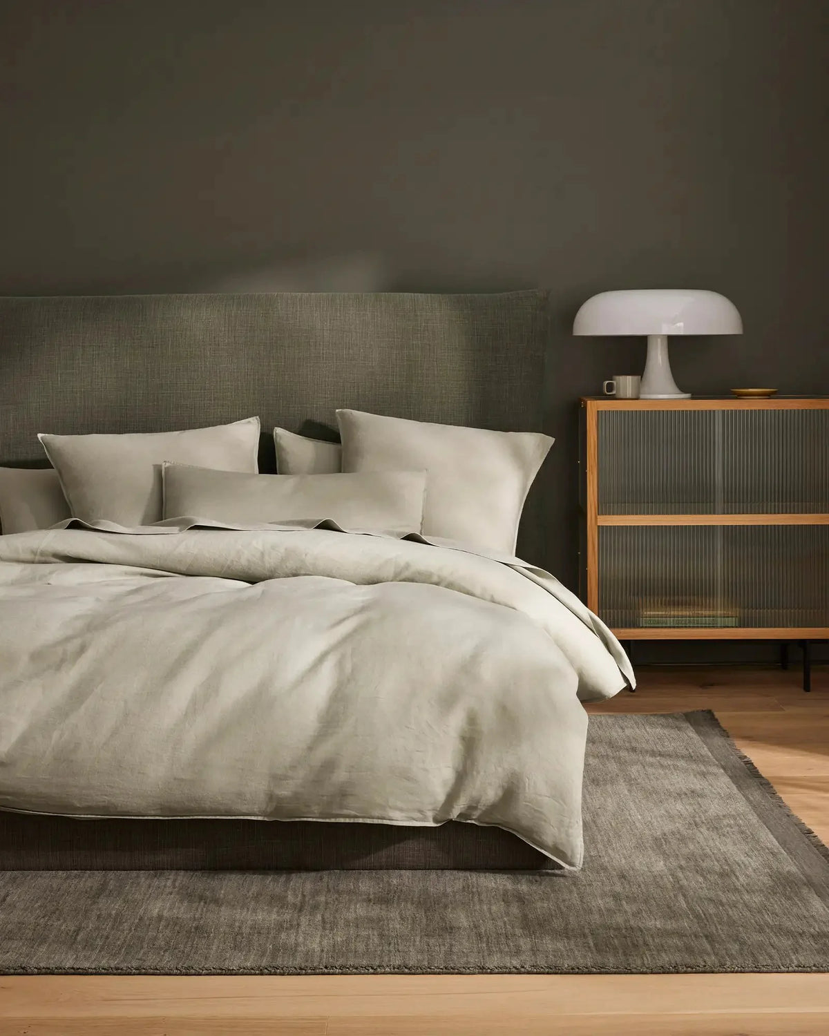 Sage green bedroom with natural linen bedding, neutral headboard, wooden mid-century nightstand, and layered textured rug in earthy tones