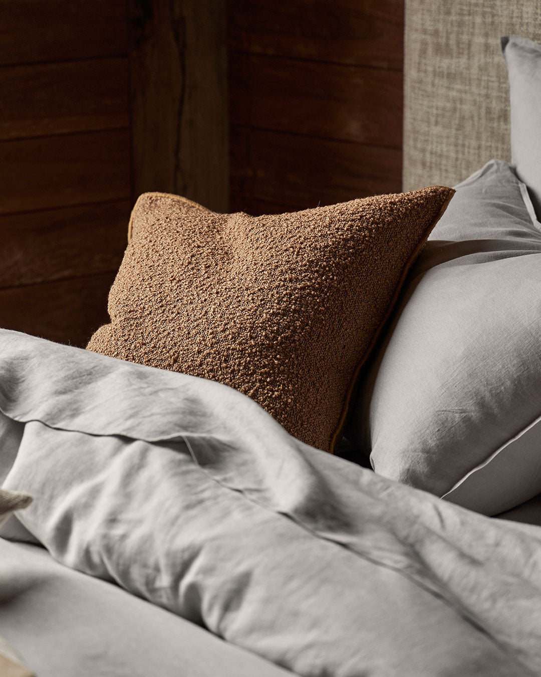 Neutral linen bedding in silver-grey tones layered with a textured caramel boucle cushion on a timber bed frame, creating warm earthy contrast and tactile bedroom styling
