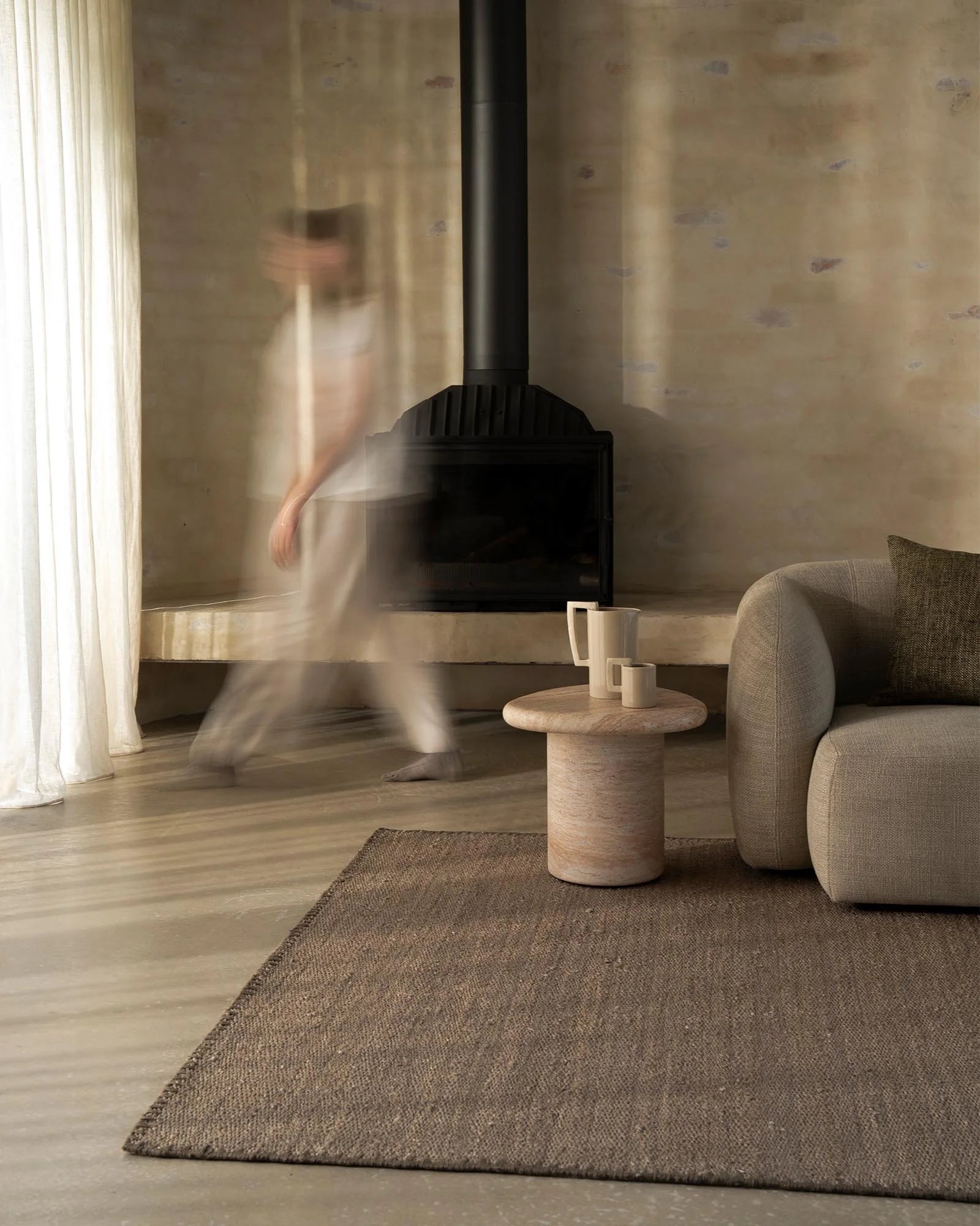 Minimalist living room with neutral taupe rug, cream linen sofa, and natural wood side table by fireplace, soft natural light from pleated curtains