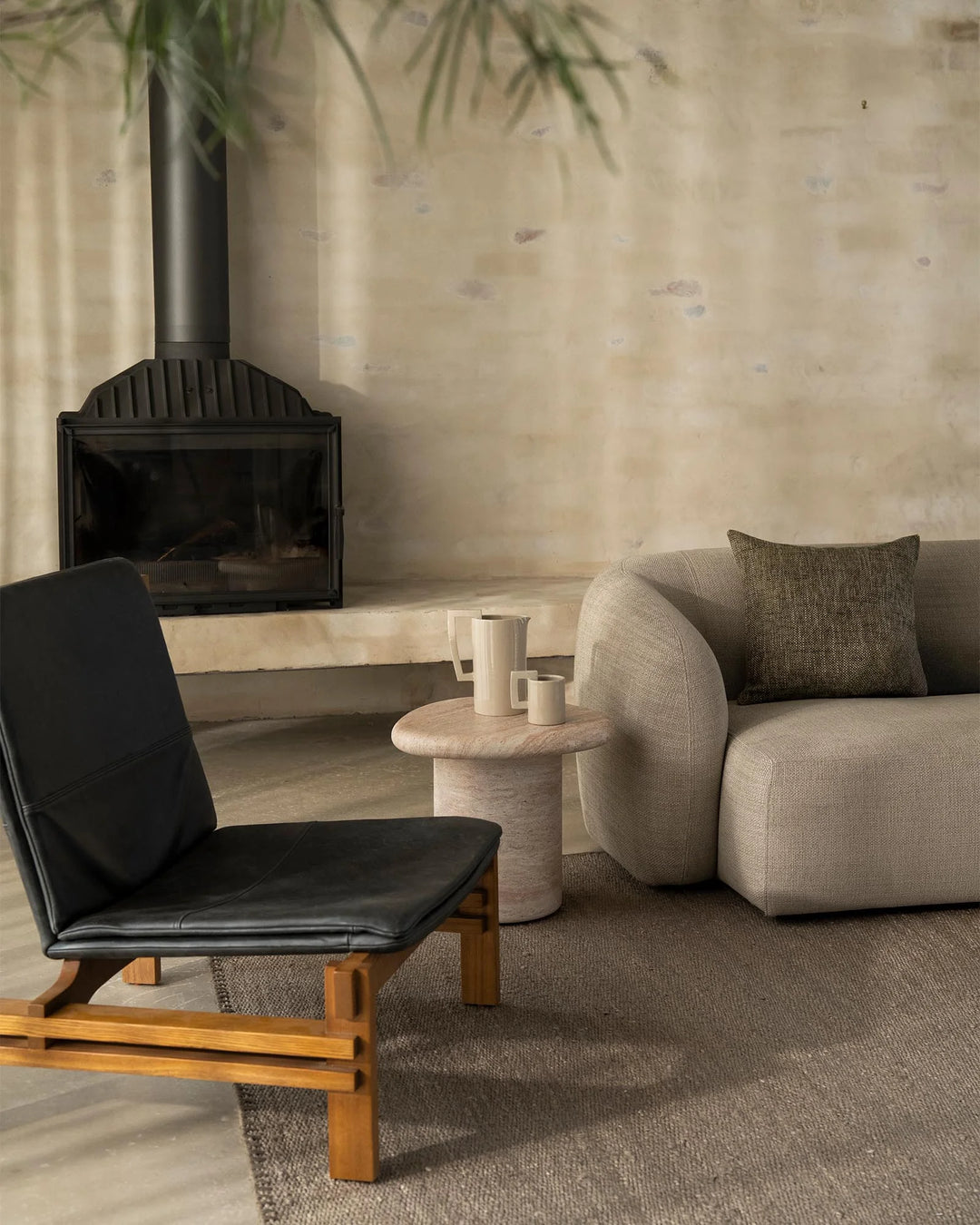 Minimalist living room with charcoal fireplace, cream curved armchair, black leather lounge chair, and soft grey-brown Monroe rug layering natural wood and neutral textures