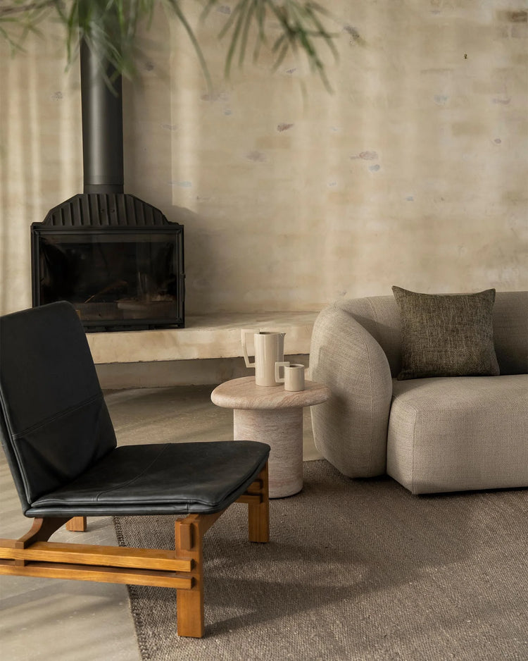 Minimalist living room with charcoal fireplace, cream curved armchair, black leather lounge chair, and soft grey-brown Monroe rug layering natural wood and neutral textures