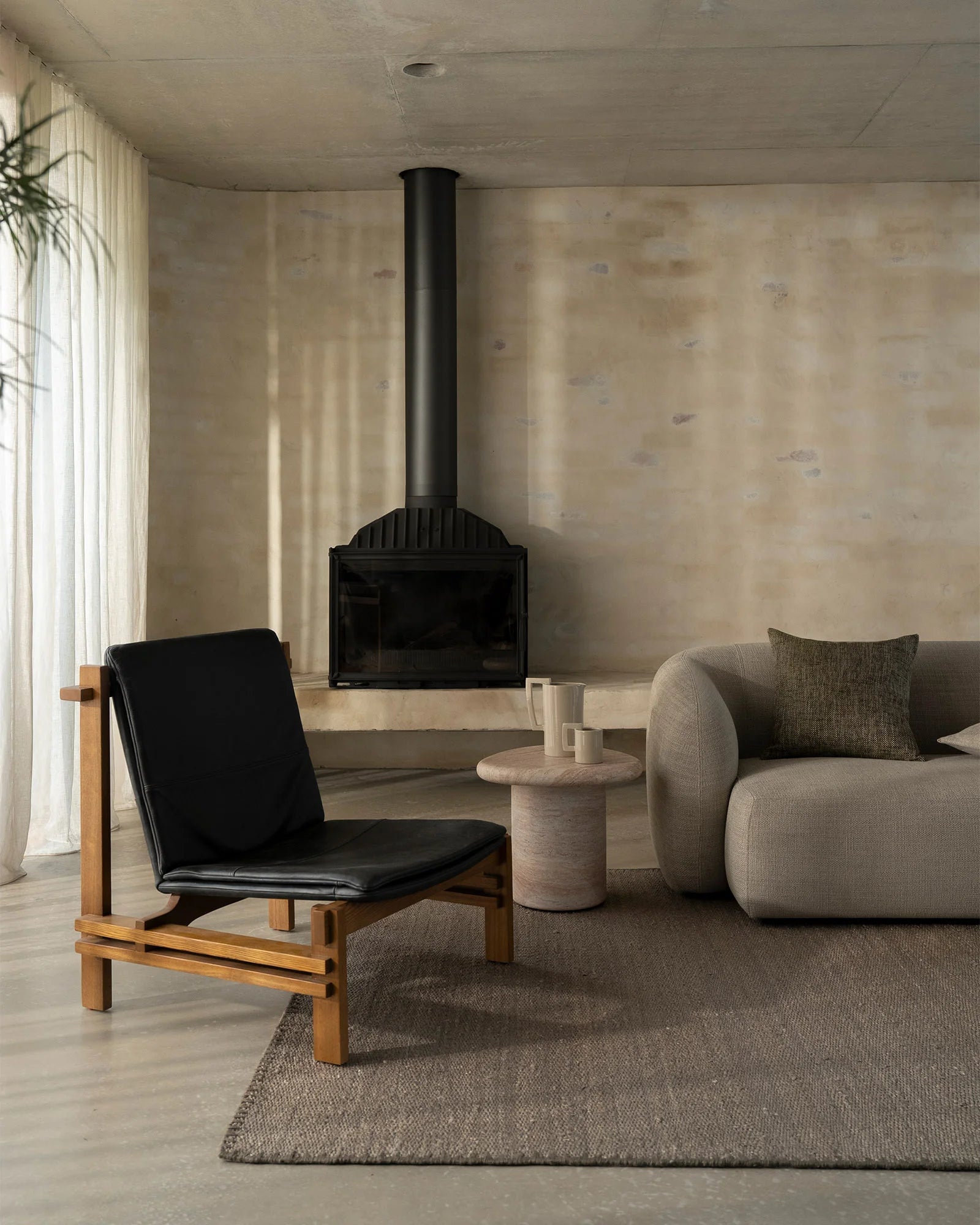 Minimalist loft interior with neutral taupe walls, black wood stove, cream curtains, and Monroe Flint rug anchoring a seating area with mid-century lounge chair and curved sofa