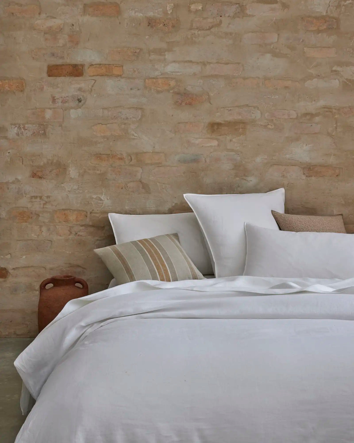Scandinavian bedroom with cream brick wall, layered white linen bedding, striped neutral cushion, and terracotta ceramic vessel styling modern minimal sleep space