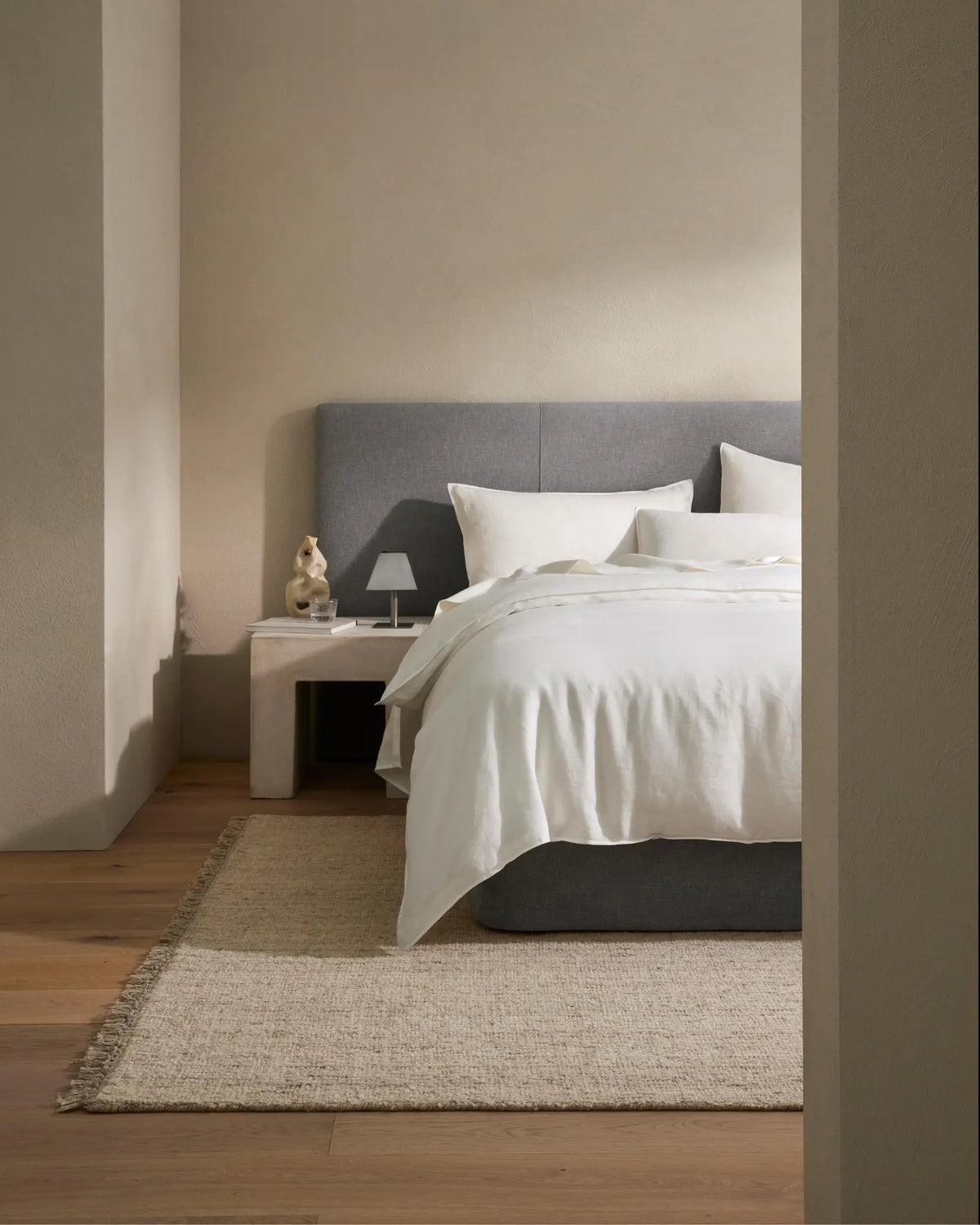 Minimalist bedroom with grey upholstered headboard, white linen bedding layered with cream pillows, natural jute rug, warm timber flooring and soft neutral wall palette.