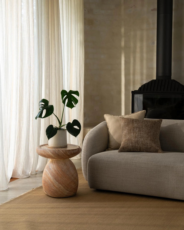 Neutral minimalist living room with warm timber side table, beige linen armchair, textured cushions and natural light filtering through cream curtains