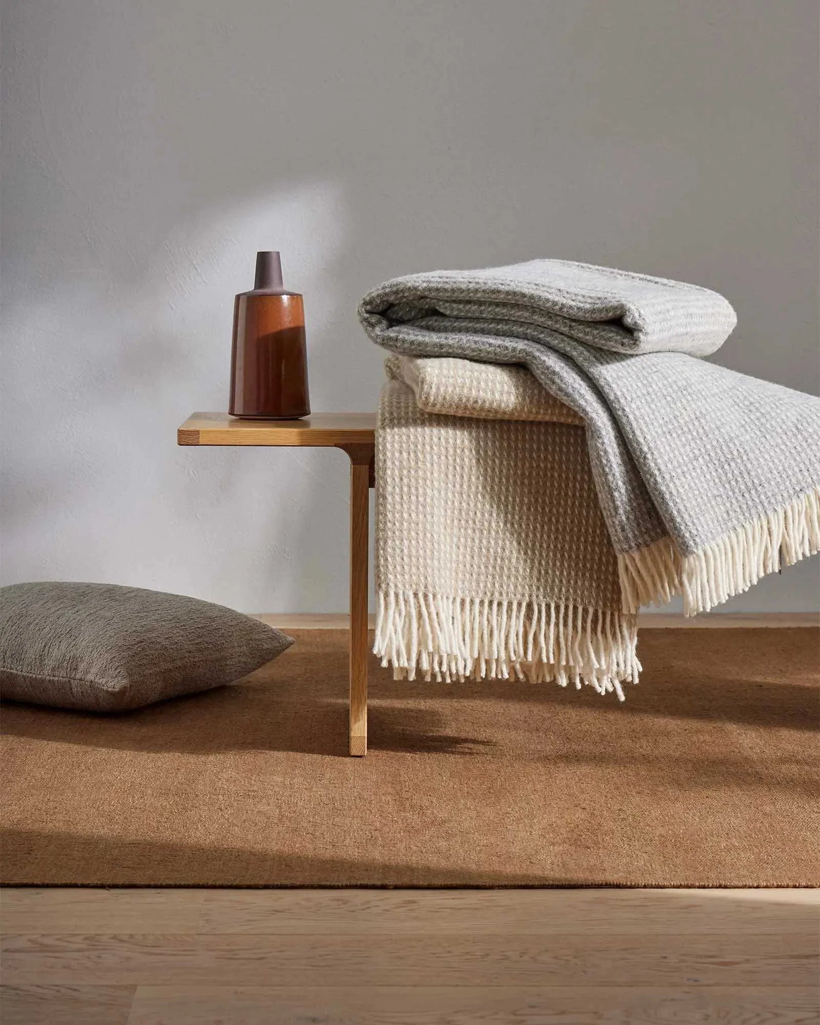Neutral toned bedroom scene with stacked throws in cream and grey wool, textured cushion, and wooden side table against soft white wall, earthy aesthetic