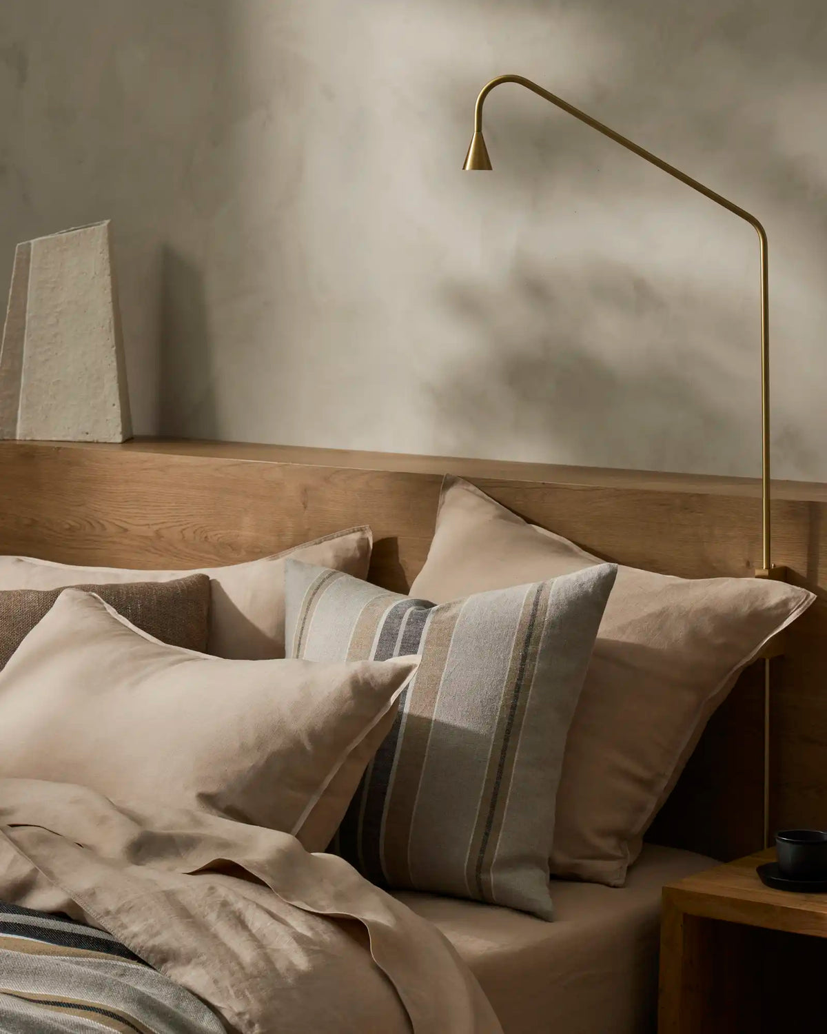 Neutral bedroom with Ravello Flat Sheet in shell, layered with striped linen pillows and taupe bedding, warm wooden bed frame, brass arc lamp, minimalist Scandinavian aesthetic