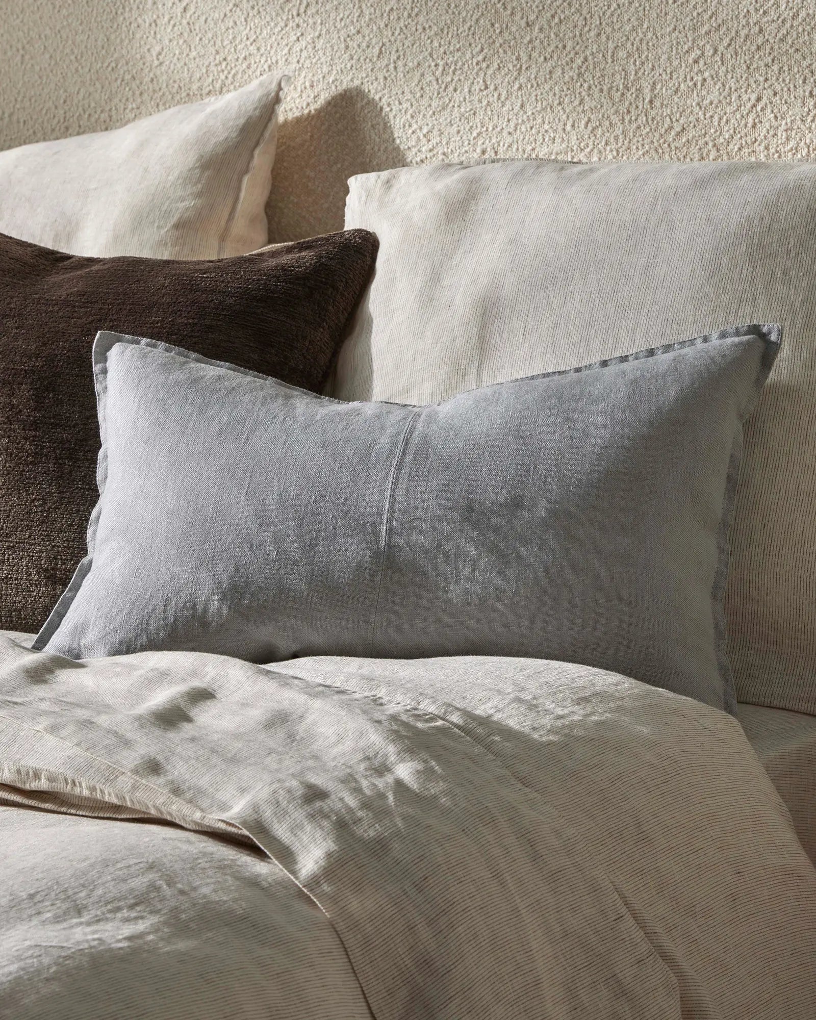 Neutral bedroom with layered pillows in cream, grey and charcoal tones, featuring textured linen and ribbed fabrics against a woven wall backdrop