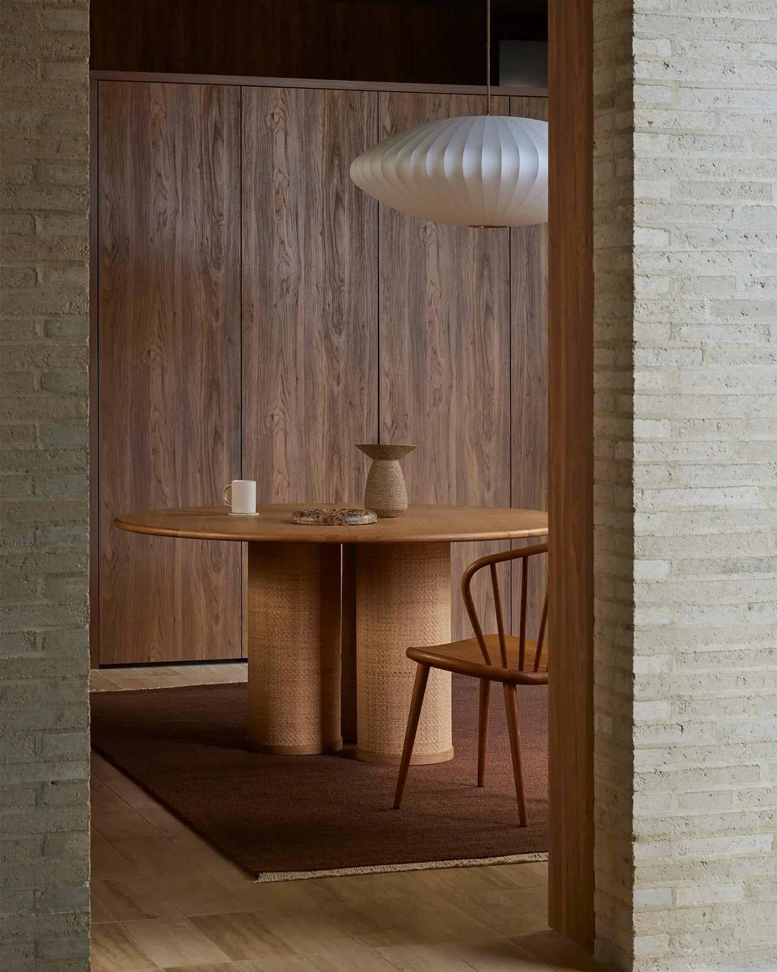 Warm timber dining nook with round wooden table, woven base, and mid-century chair beneath sculptural pendant, styled on terracotta wool rug against exposed brick backdrop
