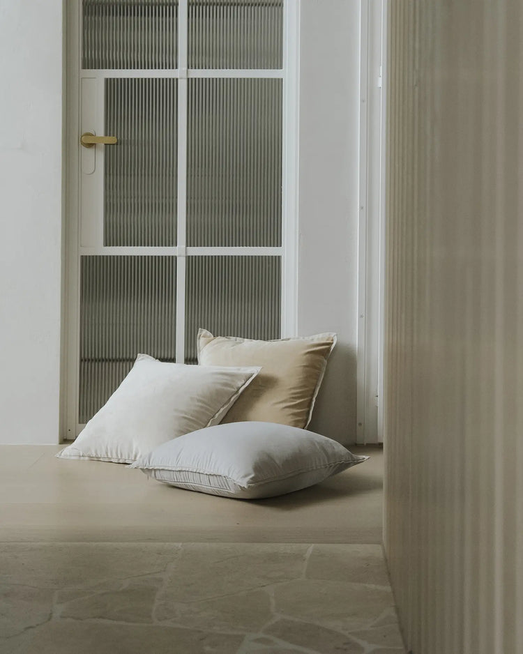 Minimalist bedroom nook with cream and sage green palette, featuring stacked Francesca cushions in frost and salt tones against ribbed glass doors and soft natural light