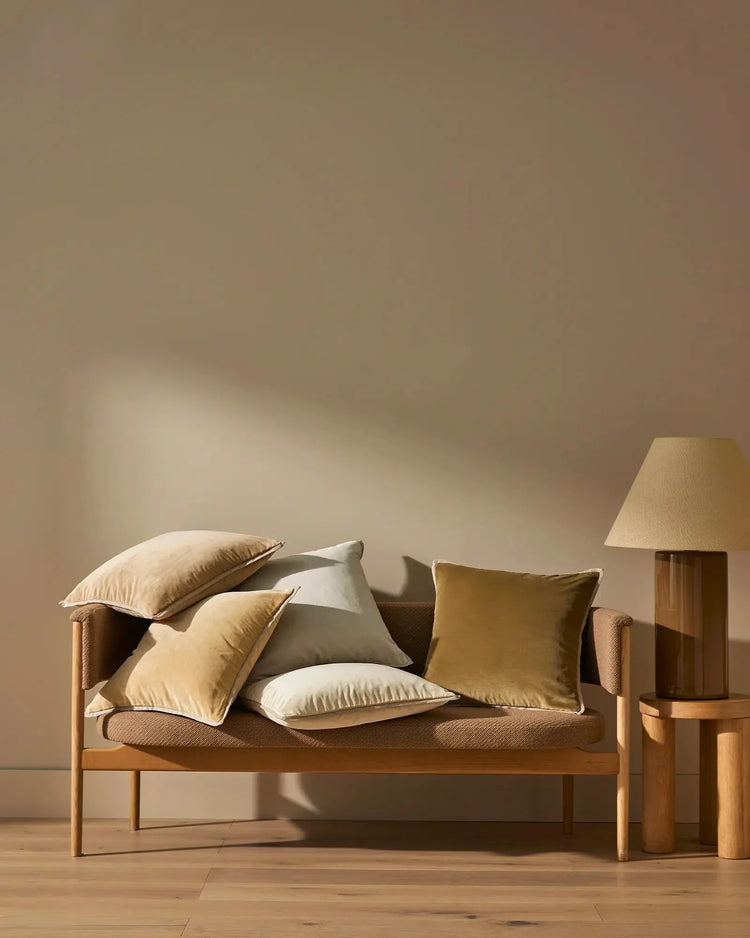 Warm minimalist living room with wooden frame sofa styled with Francesca cushions in cream and olive tones, complemented by natural wood side table and brass lamp against neutral taupe walls