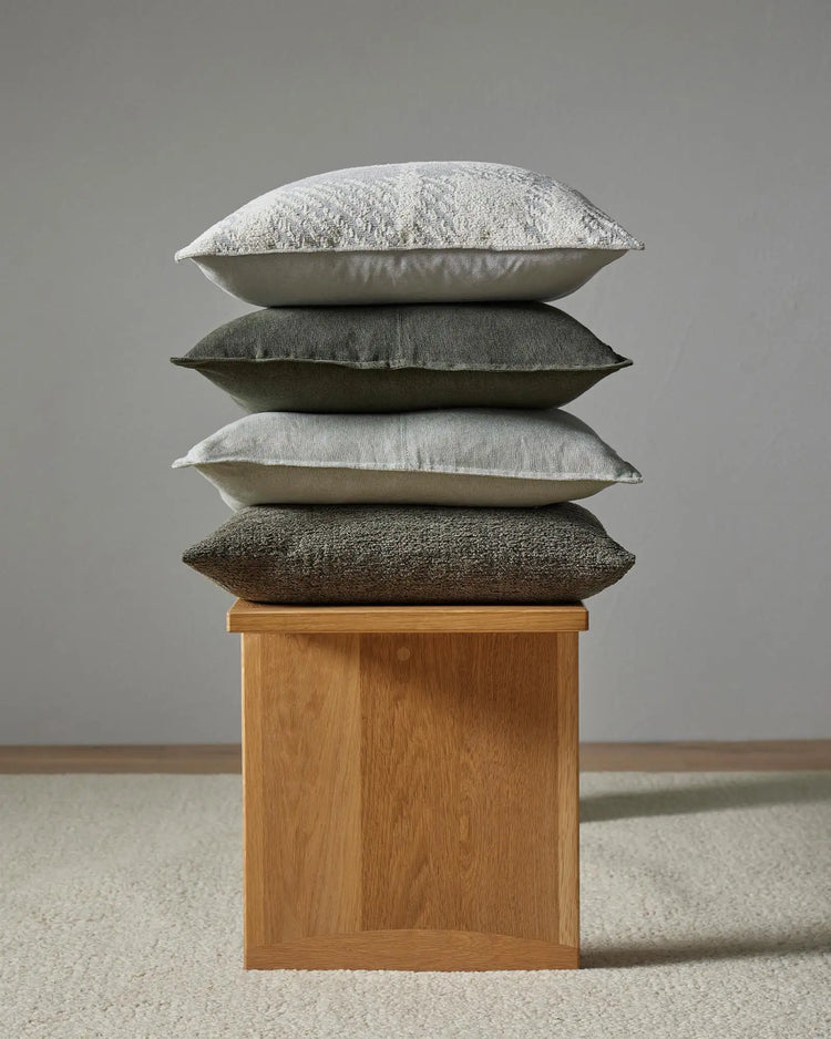 Stacked neutral cushions in cream, sage and charcoal tones showcase layered textures from linen and cotton blends, styled on a timber block base against soft grey backdrop