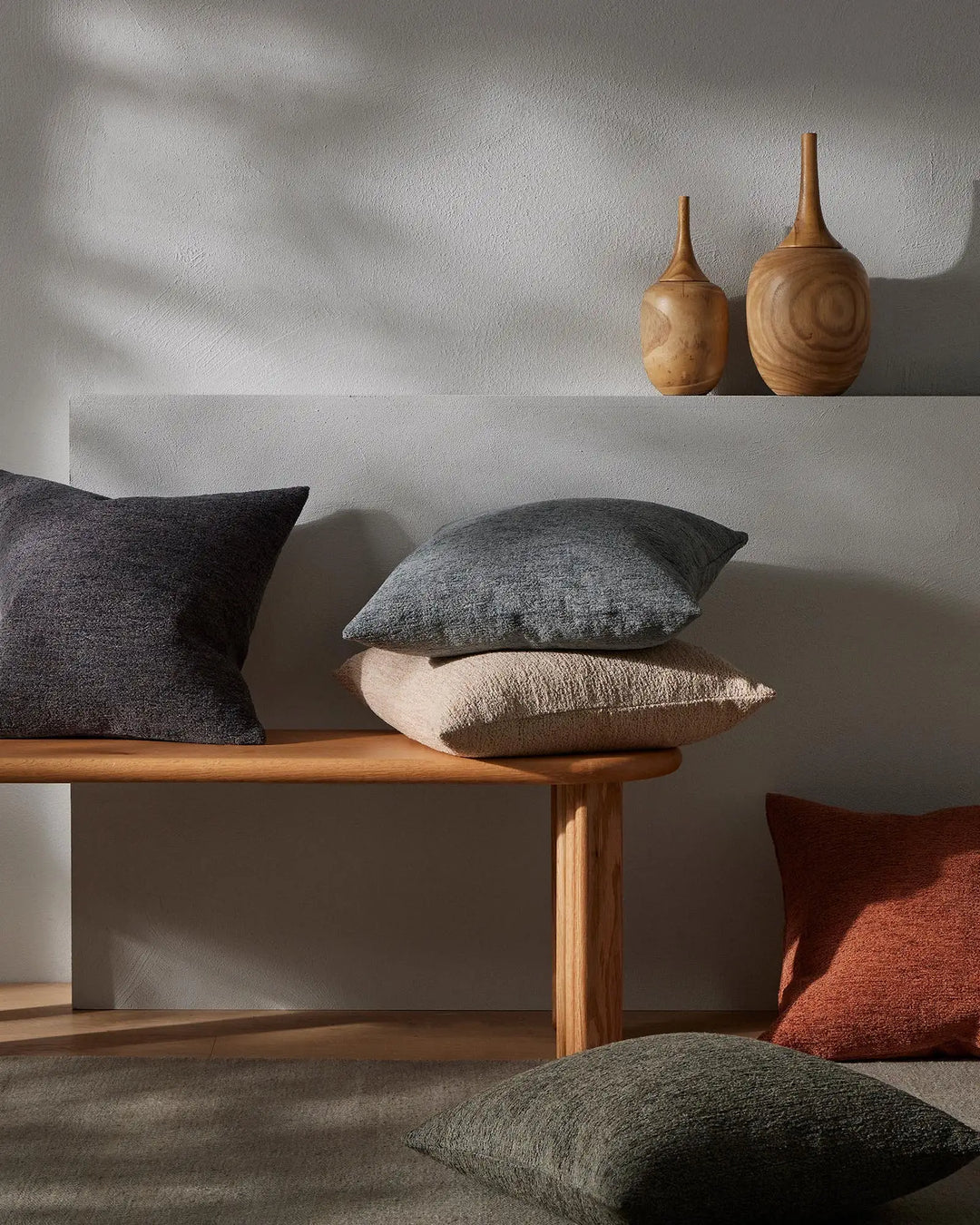 Neutral minimalist interior with Laurent cushions in charcoal, slate grey and terracotta tones layered on timber bench, natural wood carved vessels above, warm earthy palette
