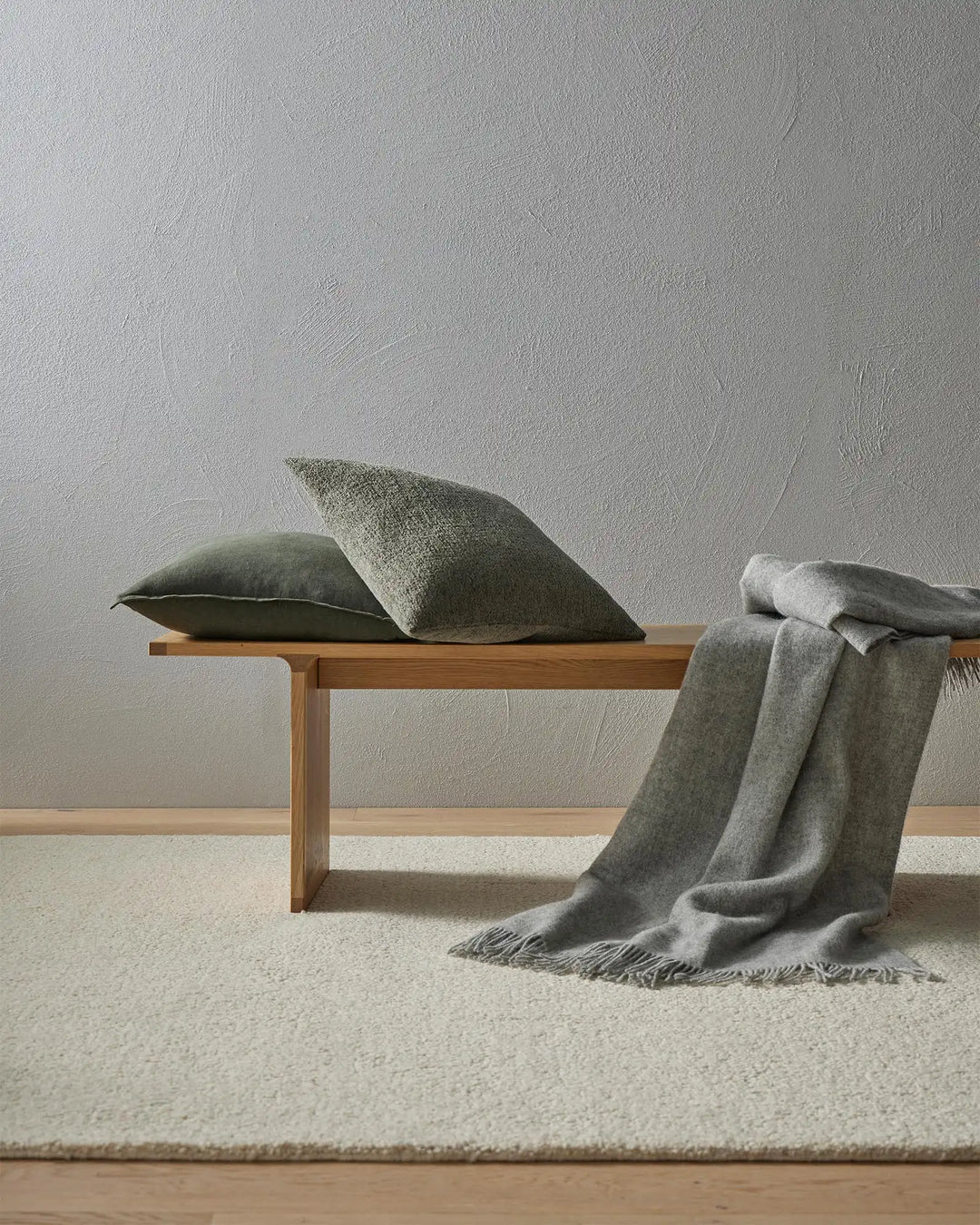 Sage green and charcoal cushions and throw draped on timber bench against textured plaster wall, warm minimalist aesthetic with natural fibres and neutral tones