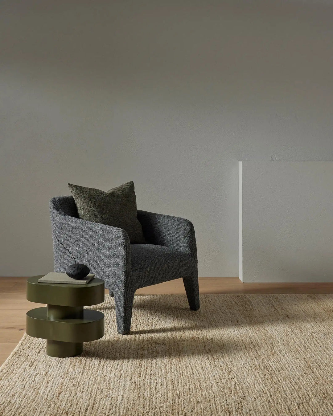 Minimalist living room with charcoal armchair and moss-green cushion on natural jute rug, sage side table, neutral walls and wood flooring