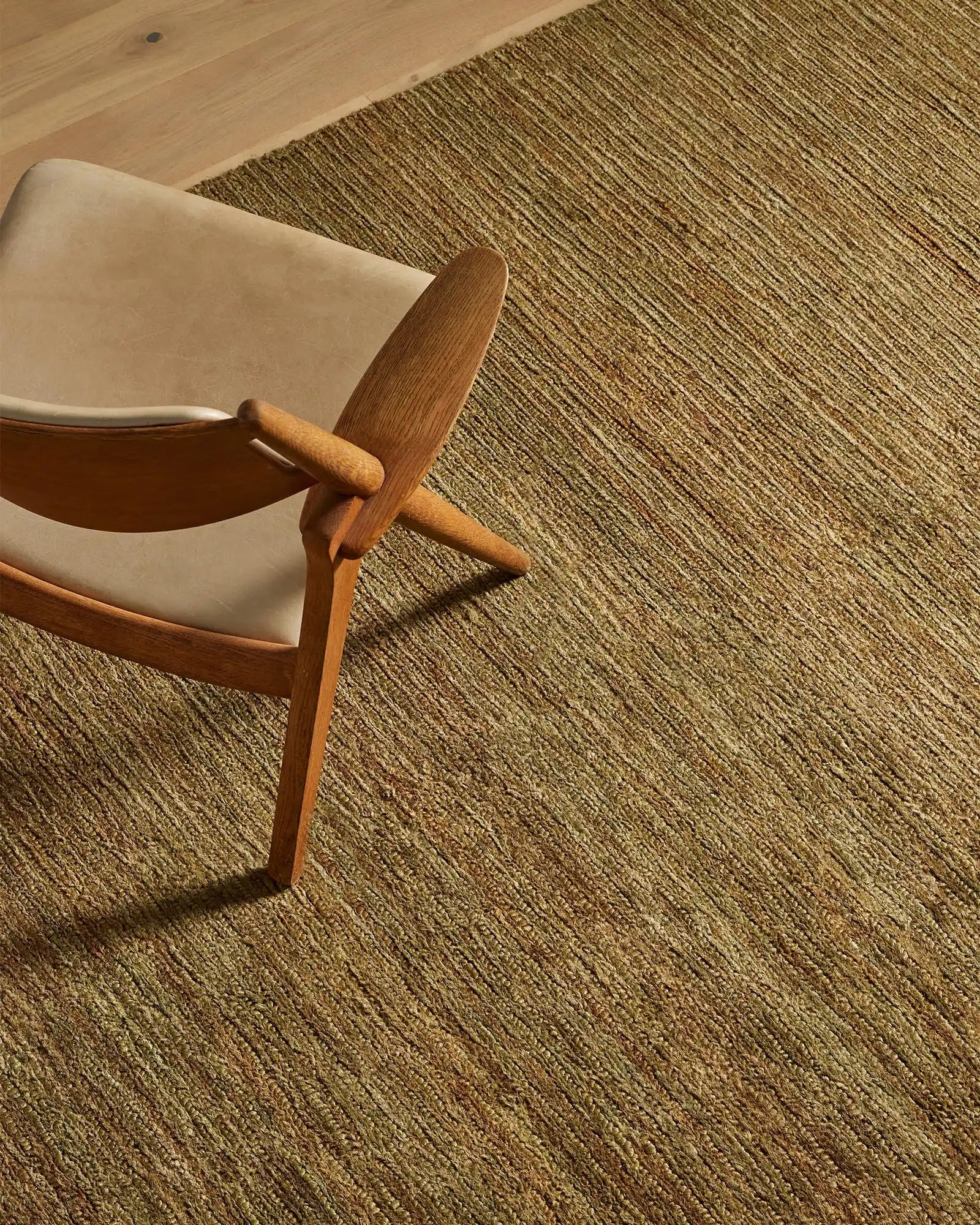 Mid-century wooden chair with cream upholstery on textured Bastian oakmoss jute rug, warm neutral palette with natural light casting shadows across woven floor covering