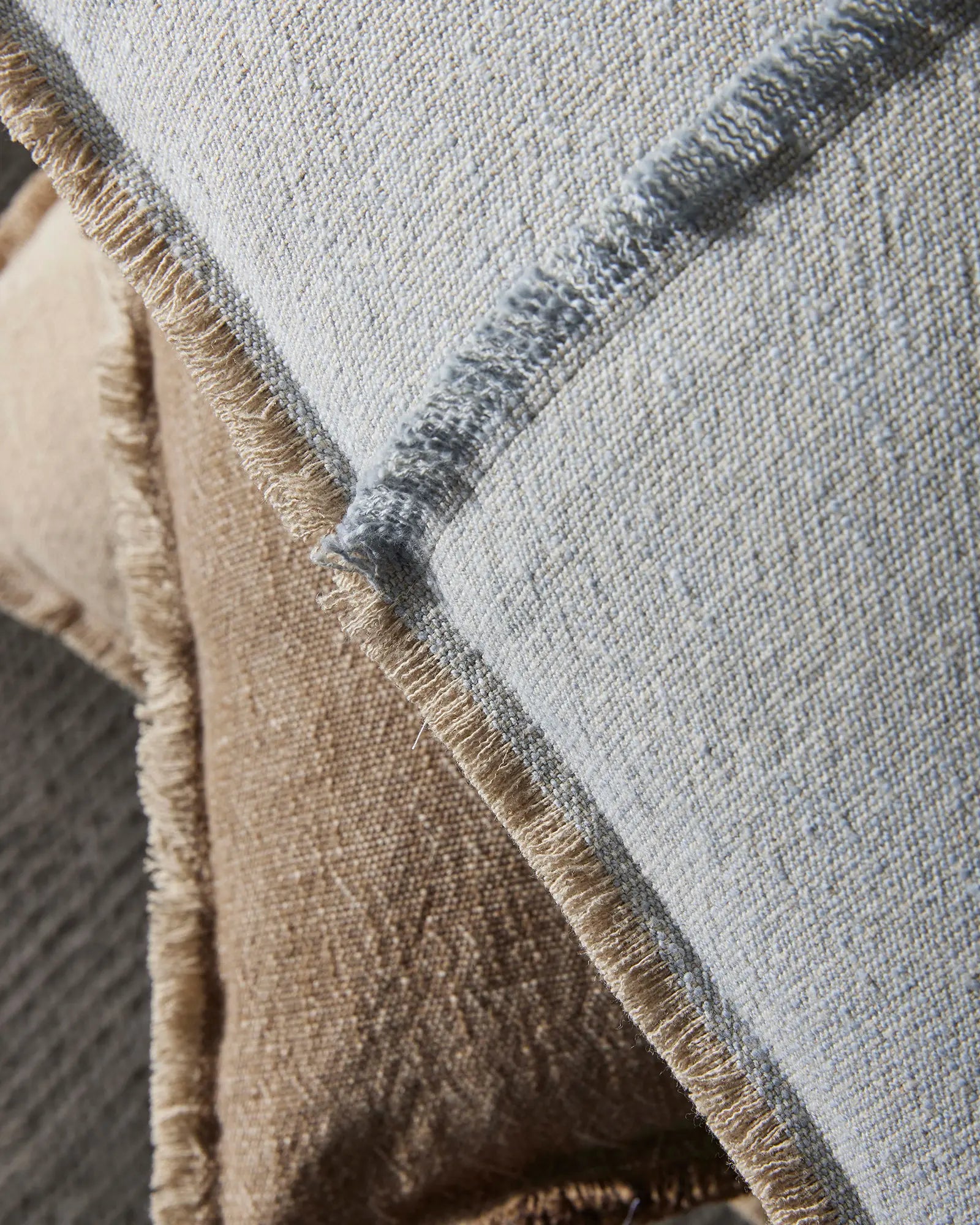 Close-up of layered cushions in neutral tones—soft grey-blue and warm taupe—with textured linen and cotton weave, fringe detailing, and natural sunlight highlighting the fabric composition