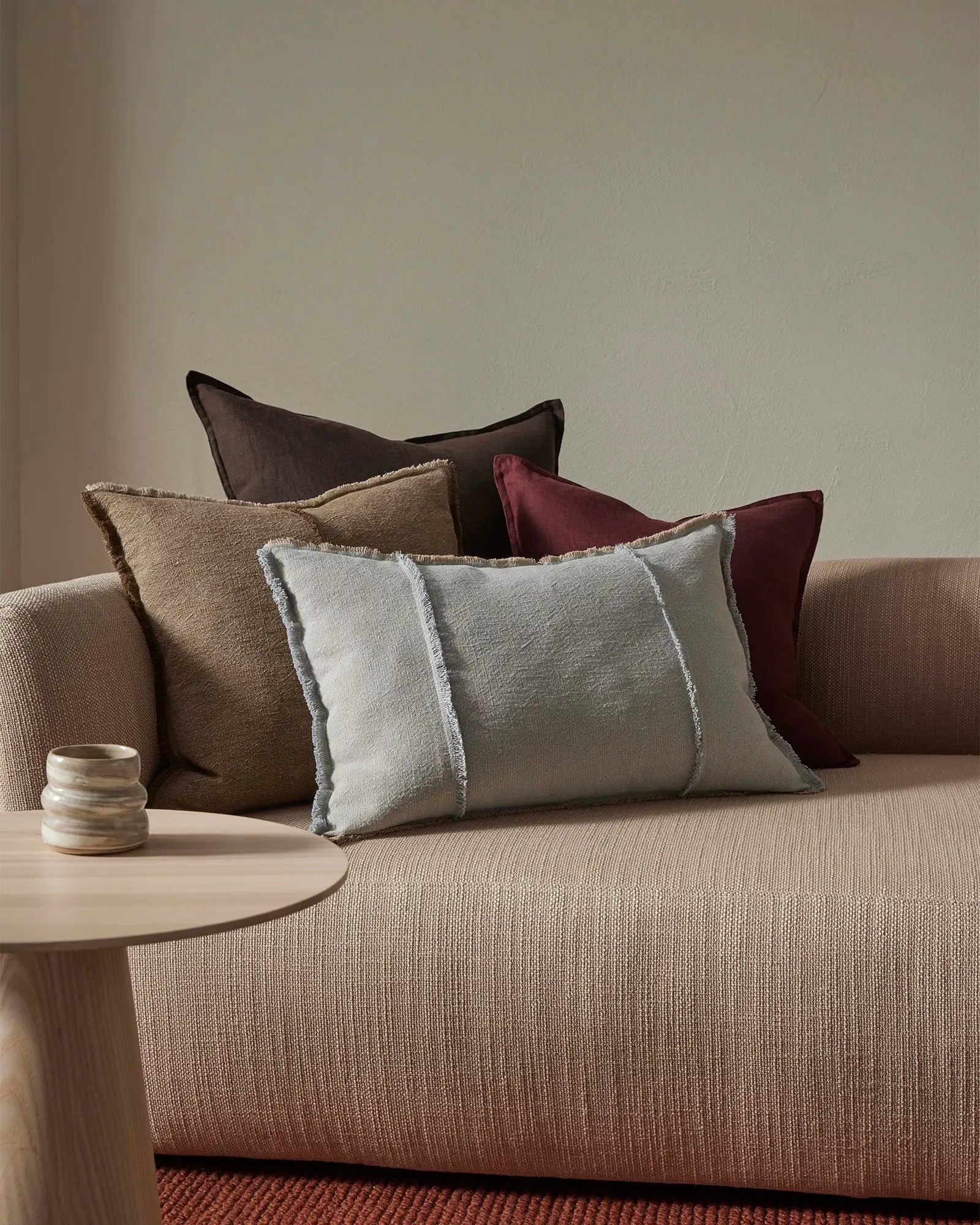Neutral living room layered with earth-tone cushions in cream, taupe, charcoal and burgundy on a beige linen sofa, with texture-rich fabrics and minimal styling against soft grey walls