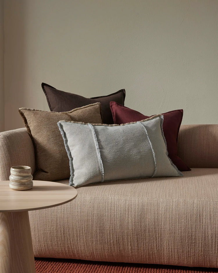 Neutral living room layered with earth-tone cushions in cream, taupe, charcoal and burgundy on a beige linen sofa, with texture-rich fabrics and minimal styling against soft grey walls