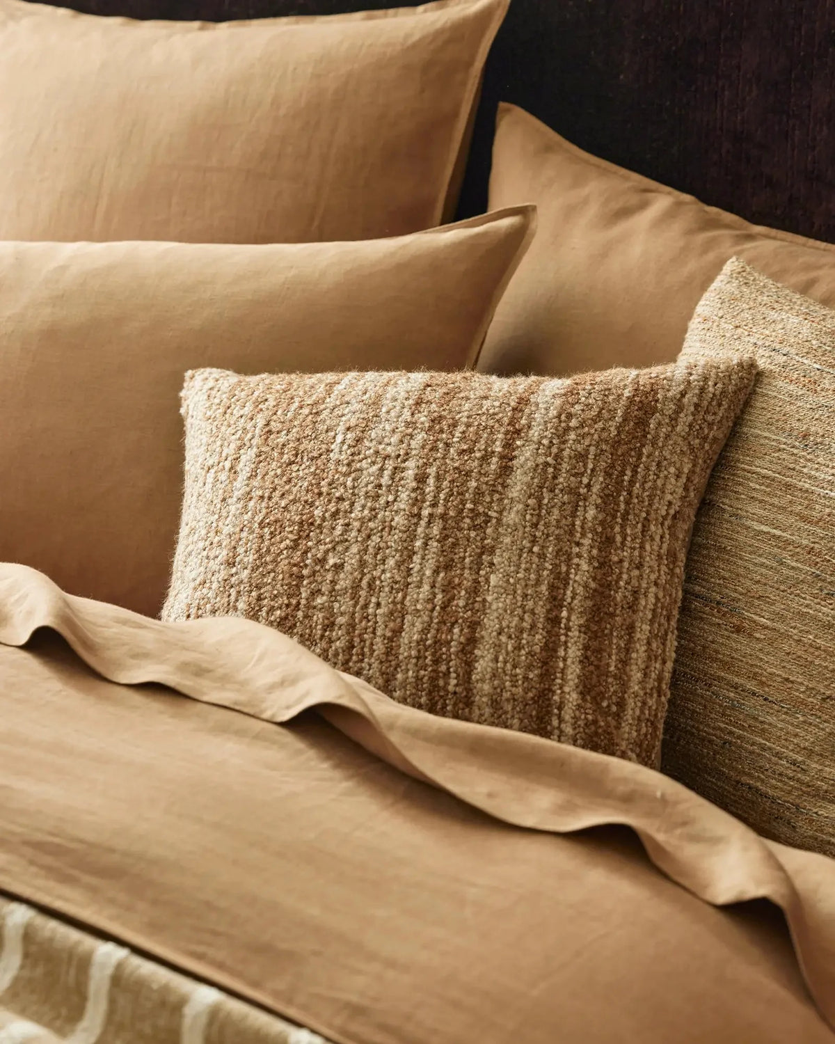 Neutral bedding styled with layered cushions in cream and natural textures, featuring crisp linen sheets and a chunky knit pillow in warm taupe tones
