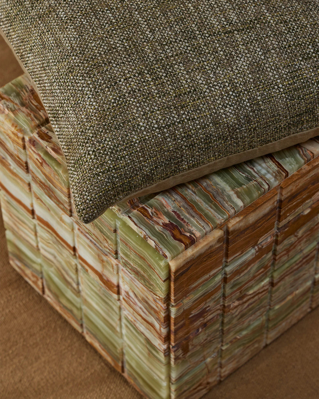 Sage-green textured cushion layered on striped upholstered ottoman with warm timber frame; earthy tones and natural fabrics create a contemporary, organic aesthetic.
