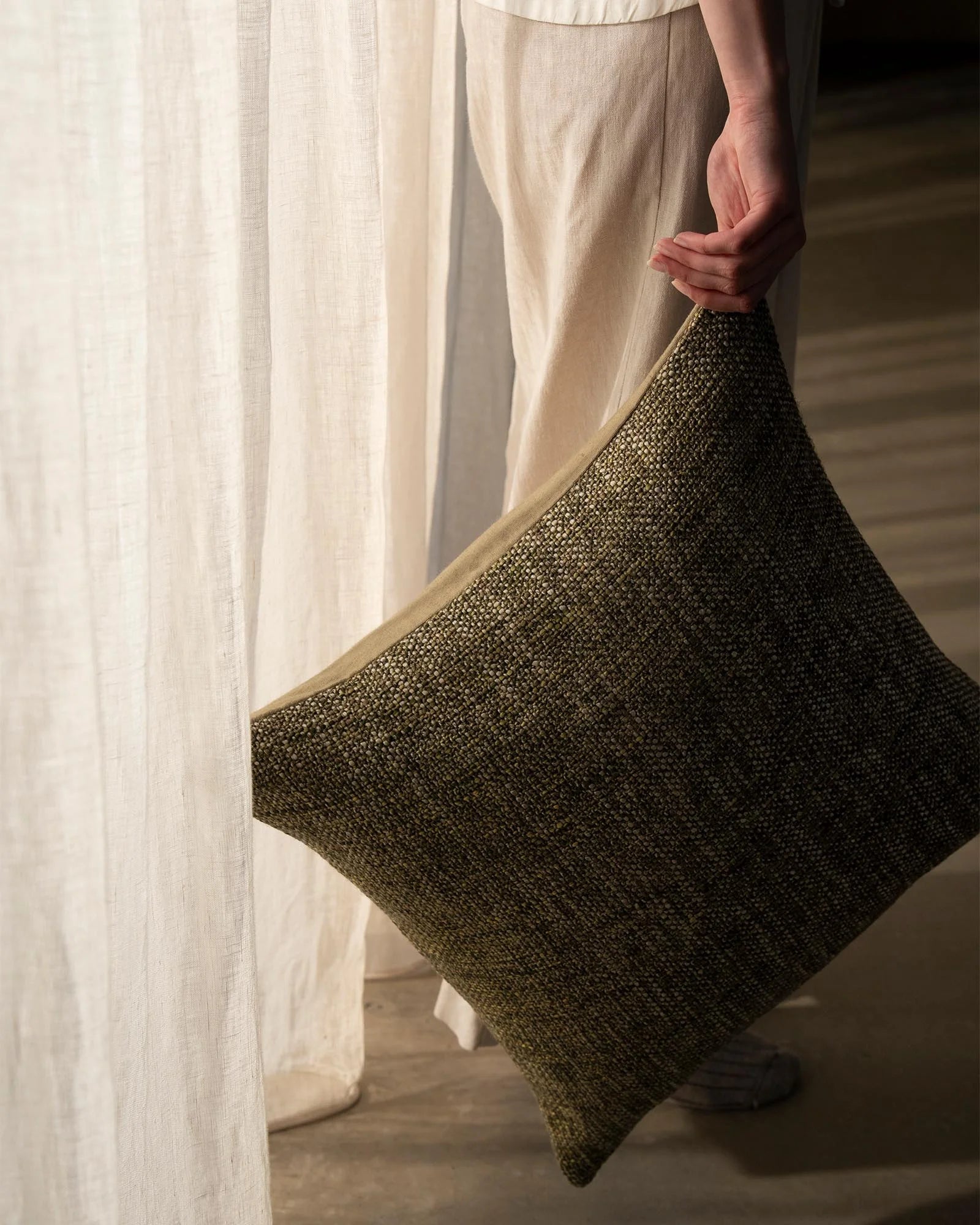 Cream linen curtains frame a charcoal textured Castel cushion in soft natural light, showcasing neutral earth-tone palette and tactile woven fabrics