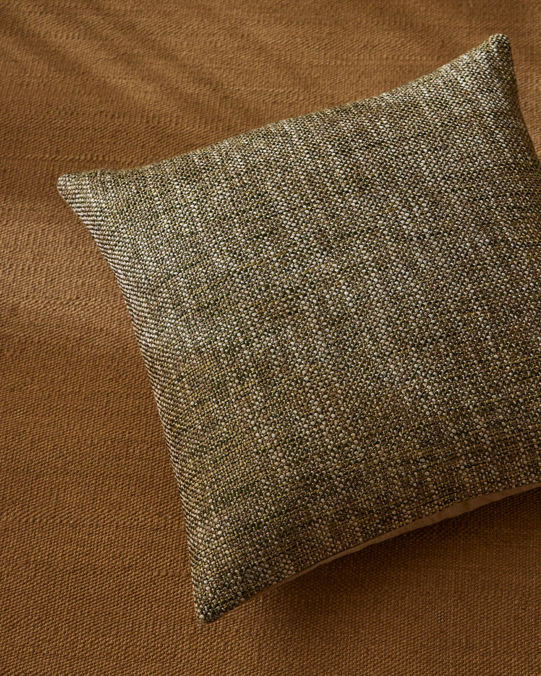 Sage-green textured cushion with woven detail layered on warm caramel wool rug, earthy neutral palette with tonal texture interplay