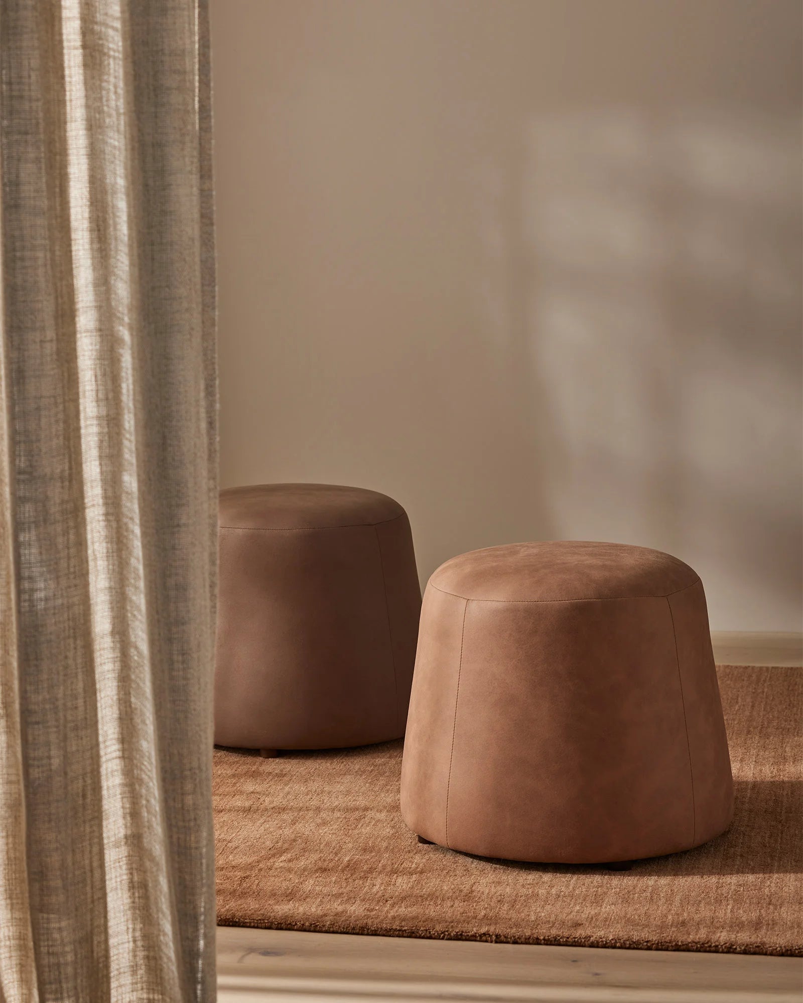 Warm neutral living room with two Landro ottomans in mocha and toffee leather, styled on natural jute rug with taupe linen curtains catching soft light
