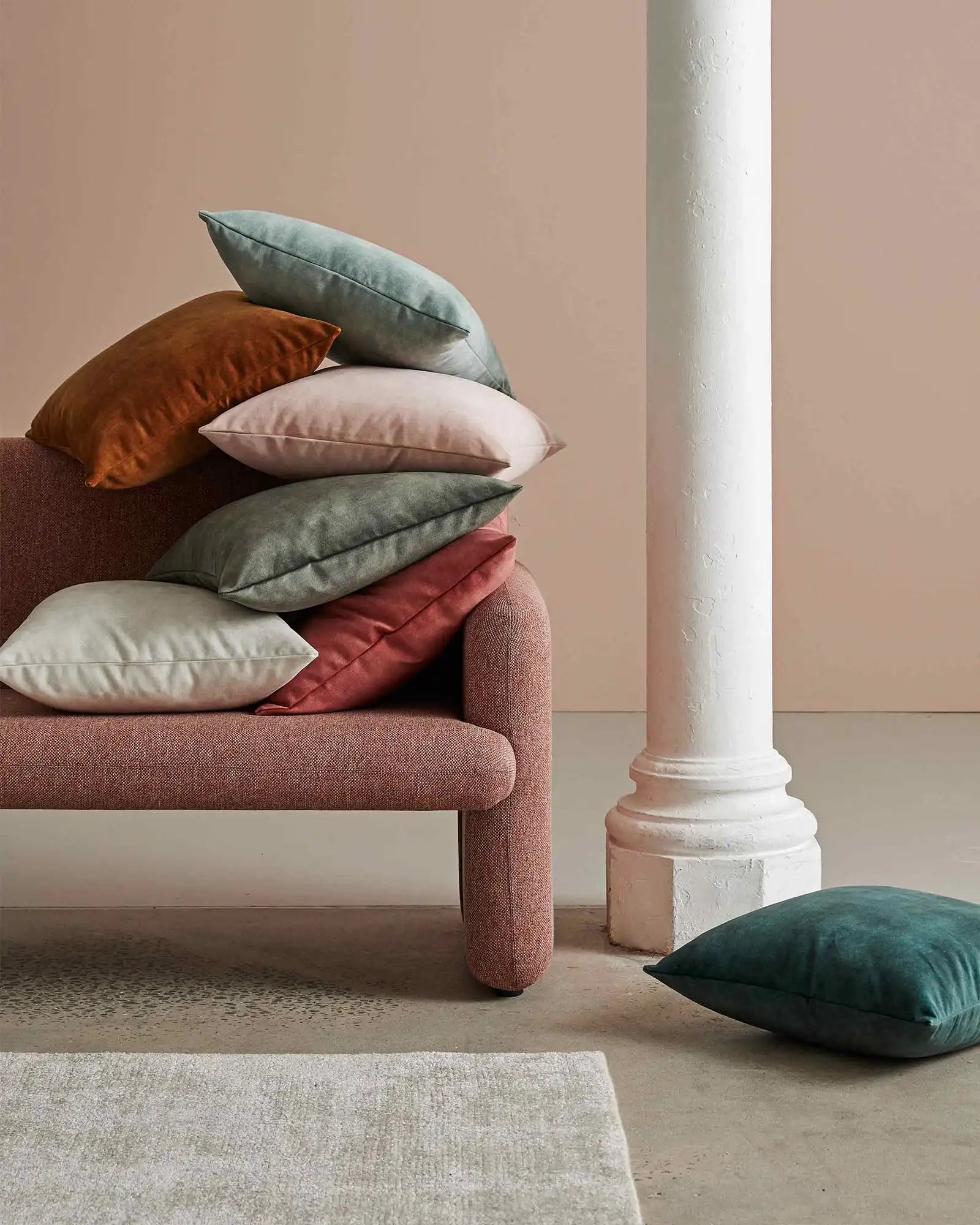 Earthy toned living room styling with stacked Ava cushions in rust, sage, cream and terracotta on a mauve upholstered bench, beside a white architectural column and neutral textured rug